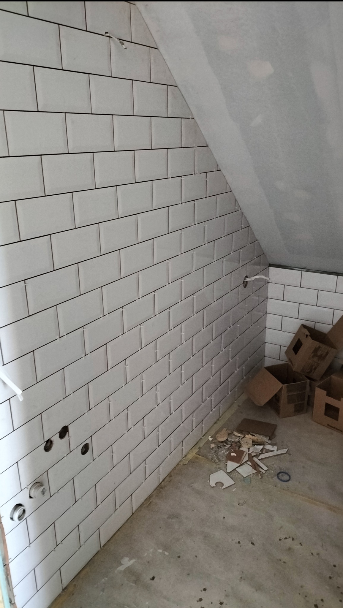 Białe, prostokątne płytki ułożone na ścianie pod skosem poddasza. Widoczne otwory na instalacje, resztki płytek i kartony na podłodze. Prace glazurnicze w trakcie.