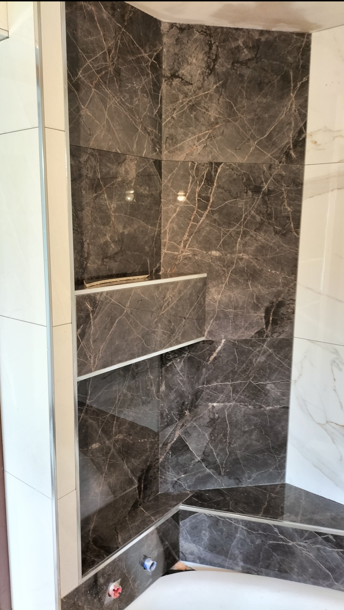 Wykończona wnęka z półkami w łazience, obłożona ciemnymi płytkami imitującymi marmur z jasnymi żyłami. Widoczne fragmenty białych płytek na ścianach.