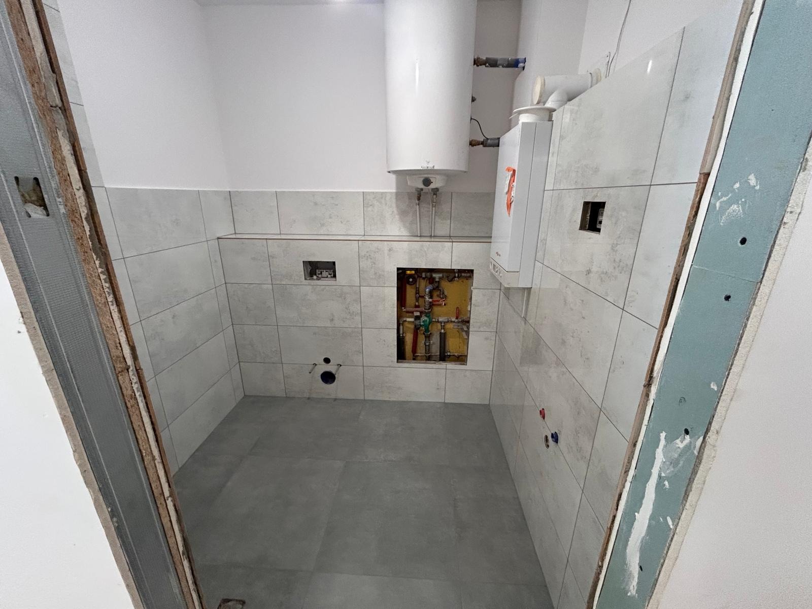 Wykończona łazienka z szarymi płytkami, widoczny bojler i piec gazowy. Otwór rewizyjny z instalacją hydrauliczną. Metalowa rama drzwiowa.