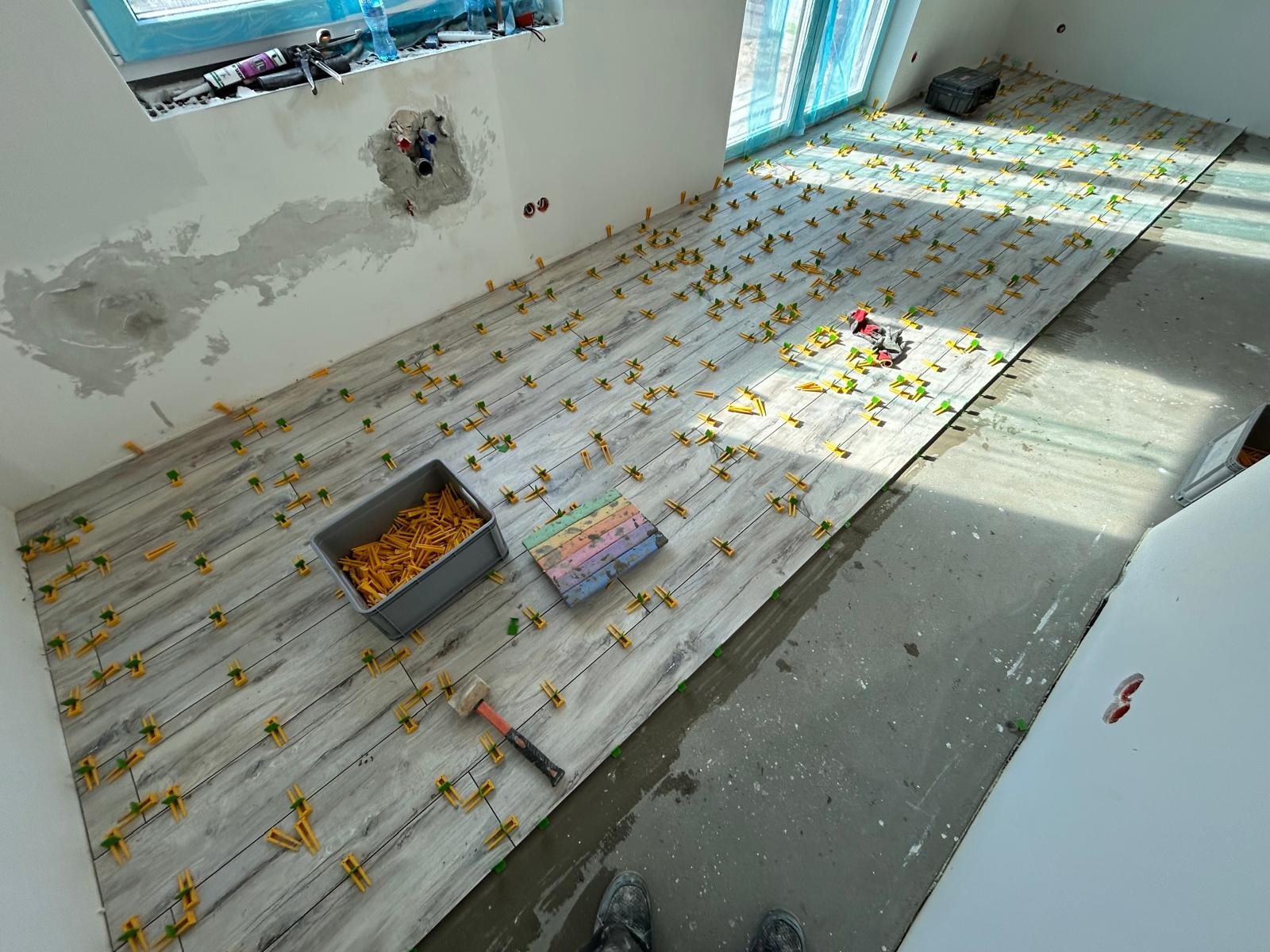 Układanie płytek podłogowych imitujących drewno, z użyciem żółtych klipsów poziomujących. Widoczny pojemnik z klipsami, młotek i poziomica. Częściowo wykończone pomieszczenie.