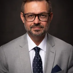 Portret mężczyzny w szarym garniturze, białej koszuli i granatowym krawacie w kropki, z okularami w rogowej oprawce na tle ciemnoszarego tła.