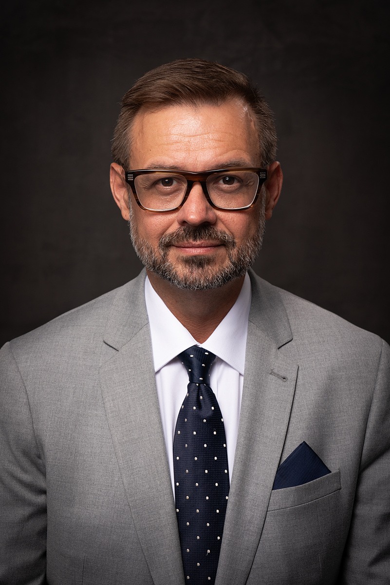 Portret mężczyzny w szarym garniturze, białej koszuli i granatowym krawacie w kropki, z okularami w rogowej oprawce na tle ciemnoszarego tła.