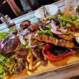 RM&KM TEAM SPÓŁKA Z OGRANICZONĄ ODPOWIEDZIALNOŚCIĄ - Drewniana deska z różnorodnymi grillowanymi mięsami, frytkami, sałatą, czerwoną cebulą i pieczoną papryką, serwowana na białym, drewnianym stole restauracyjnym.