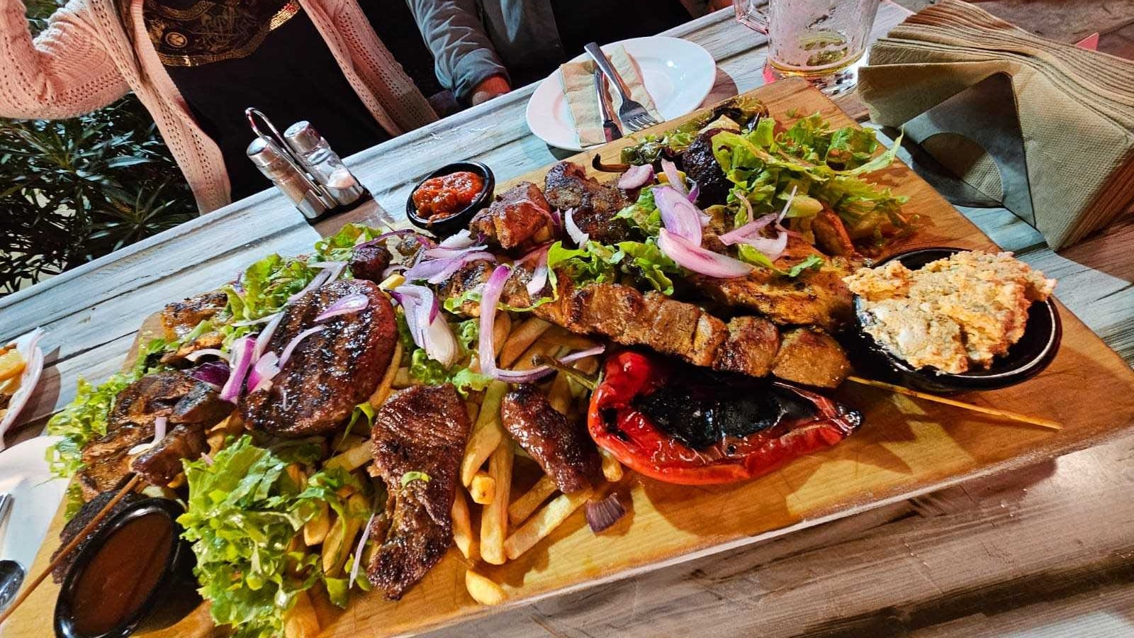 Drewniana deska z różnorodnymi grillowanymi mięsami, frytkami, sałatą, czerwoną cebulą i pieczoną papryką, serwowana na białym, drewnianym stole restauracyjnym.