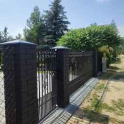 Elegancka, czarna brama wjazdowa z kutymi elementami, osadzona na murowanych słupkach z ciemnej cegły. Widoczne drzewa i fragment drogi dojazdowej.