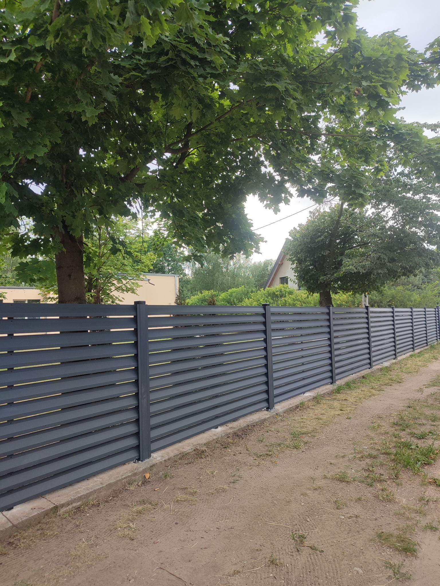 Nowoczesne, poziome ogrodzenie panelowe w kolorze grafitowym, z widocznym betonowym podmurówką, na tle zieleni drzew i zabudowań. Ujęcie z poziomu gruntu.