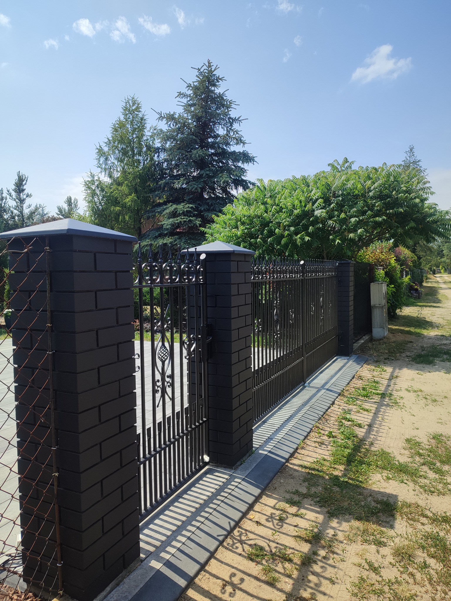 Elegancka, czarna brama wjazdowa z kutymi elementami, osadzona na murowanych słupkach z ciemnej cegły. Widoczne drzewa i fragment drogi dojazdowej.