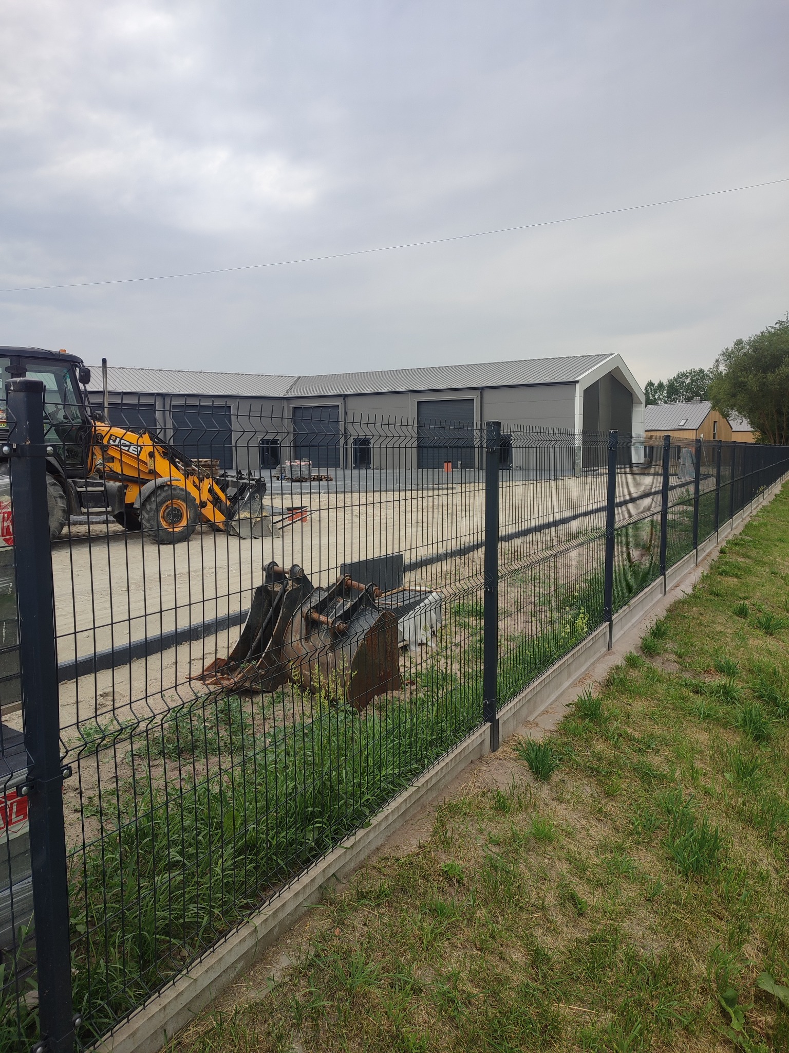 Nowoczesne ogrodzenie panelowe z podmurówką betonową, w tle budynek z bramami garażowymi i koparka. Widok na posesję.
