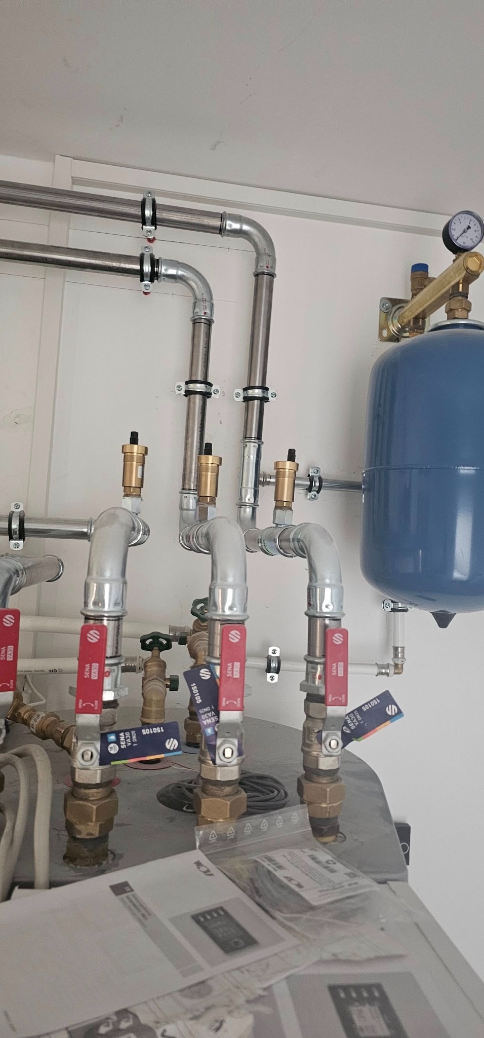 Instalacja grzewcza z widocznymi rurami, zaworami z czerwonymi dźwigniami, manometrem, zbiornikiem wyrównawczym i dokumentacją techniczną na pierwszym planie.