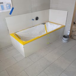 MXG Usługi Remontowe Marek Głomski - Wanna w obudowie z płyt, zabezpieczona żółtą taśmą, z zielonymi krzyżykami dystansowymi. Na podłodze szare płytki. Na ścianie widoczny projekt łazienki.