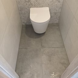 MXG Usługi Remontowe Marek Głomski - Biała, podwieszana toaleta w wykończonej łazience. Szare płytki podłogowe i dekoracyjne płytki ścienne. Fragment drewnianej ramy drzwi w dolnej części kadru.