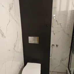 Nowoczesna toaleta podwieszana z czarną zabudową i marmurowymi płytkami. Widoczny fragment kabiny prysznicowej i gniazdko elektryczne.