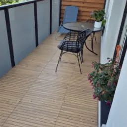 Balkon z drewnopodobną podłogą z płytek, stolikiem i krzesłami, ozdobiony zielenią w doniczkach. Widok na zewnątrz przez przeszkloną balustradę.
