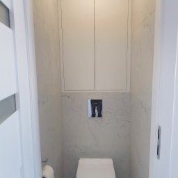 MXG Usługi Remontowe Marek Głomski - Nowoczesna toaleta w małej łazience wykończonej białymi płytkami imitującymi marmur, z zabudową nad stelażem i minimalistycznym przyciskiem spłukującym.