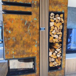 MXG Usługi Remontowe Marek Głomski - Rustykalny kominek z wnęką na drewno, wykończony metaliczną farbą z efektem rdzy i drewnianymi listwami. Nowoczesny design z elementami industrialnymi.