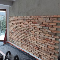 MXG Usługi Remontowe Marek Głomski - Ceglana ściana dekoracyjna w trakcie montażu, widoczne narzędzia i przygotowane podłoże. Częściowo wykończona ściana z cegły w pomieszczeniu.