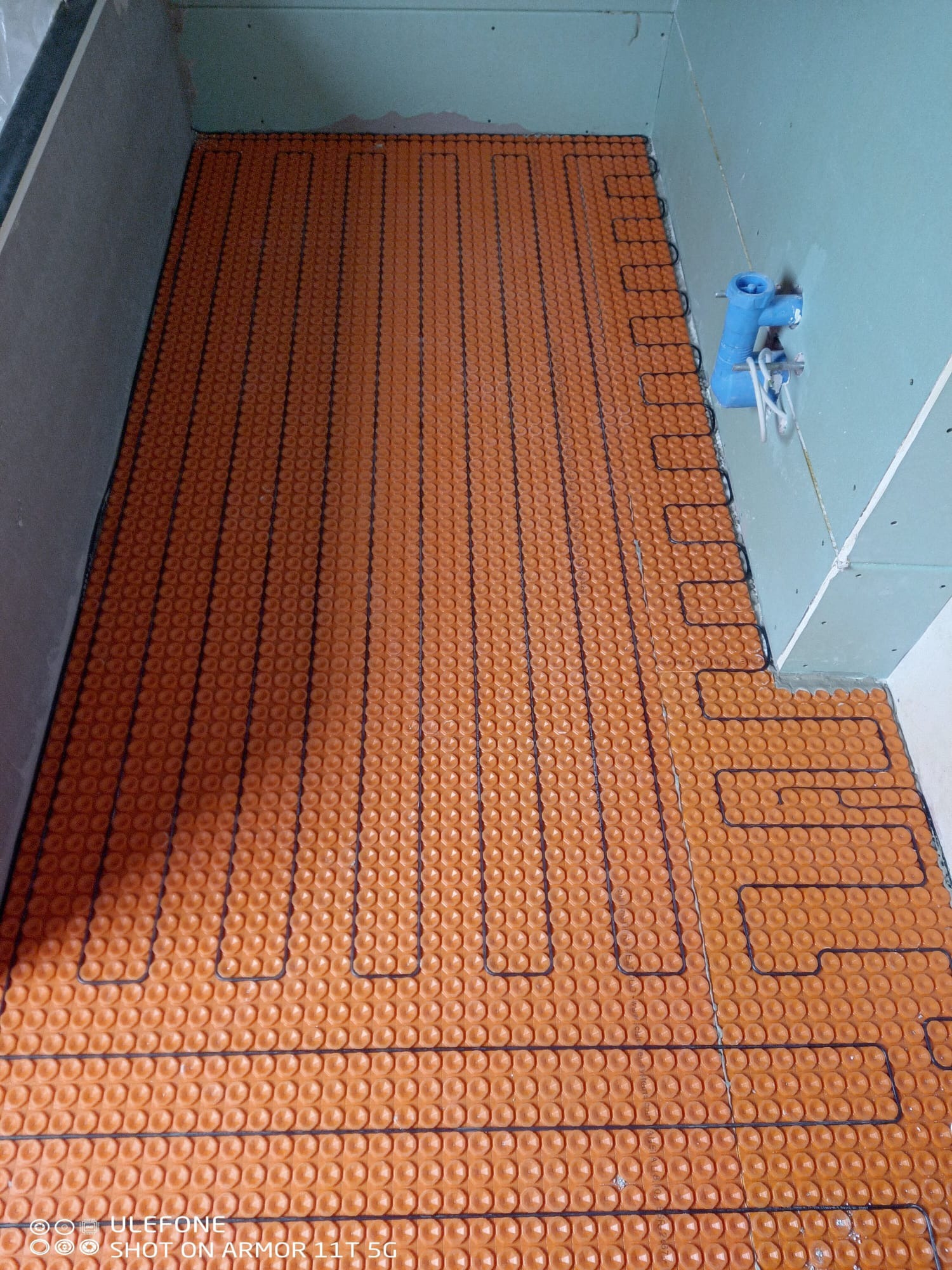 Pomarańczowa mata grzewcza z czarnymi przewodami, ułożona w surowej łazience przed wylewką. Widoczna instalacja wodna na ścianie.