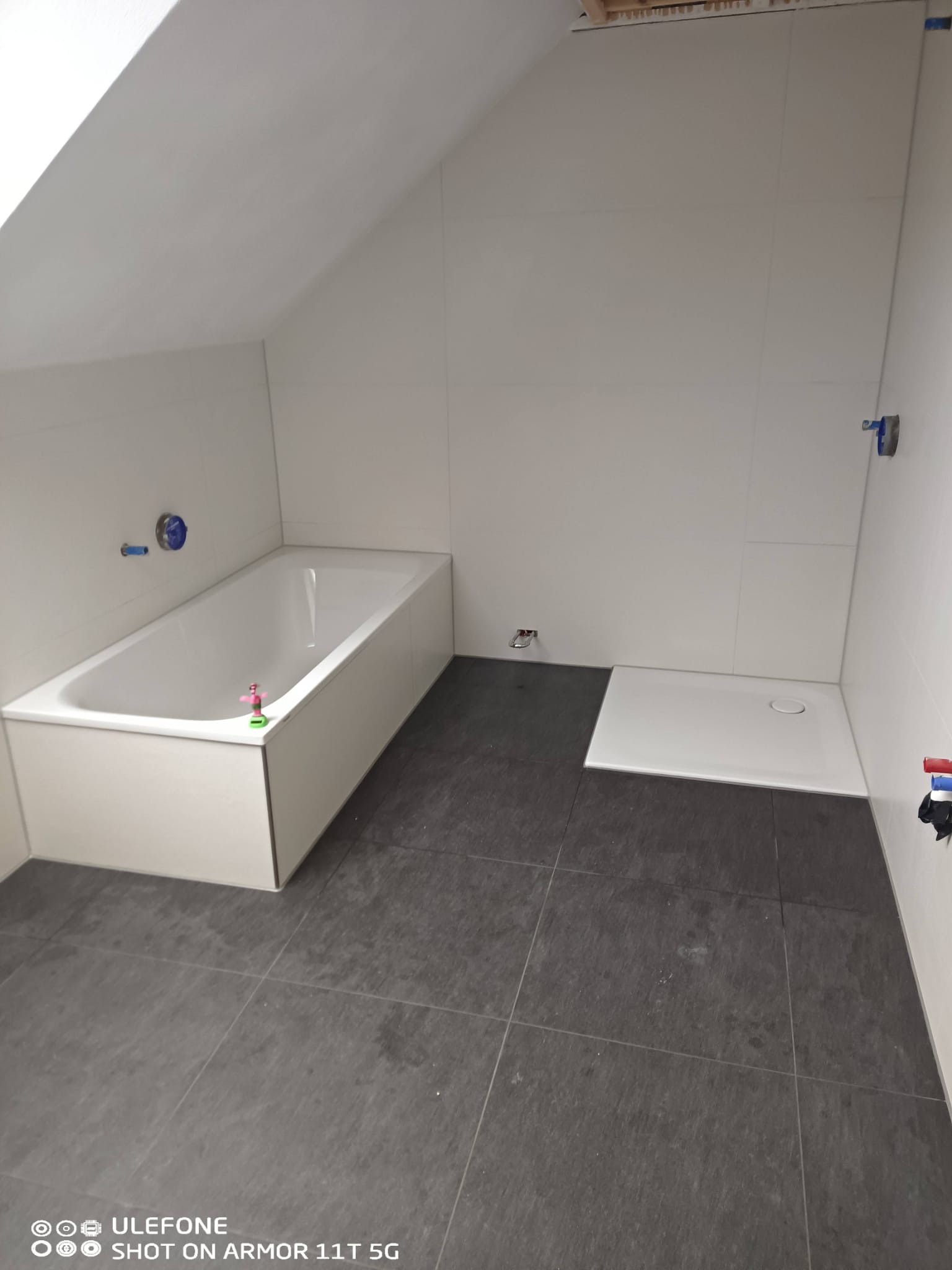 Biała wanna i brodzik w łazience na poddaszu, wyłożonej szarymi płytkami. Widoczne przyłącza wodne i fragment drewnianej konstrukcji dachu.