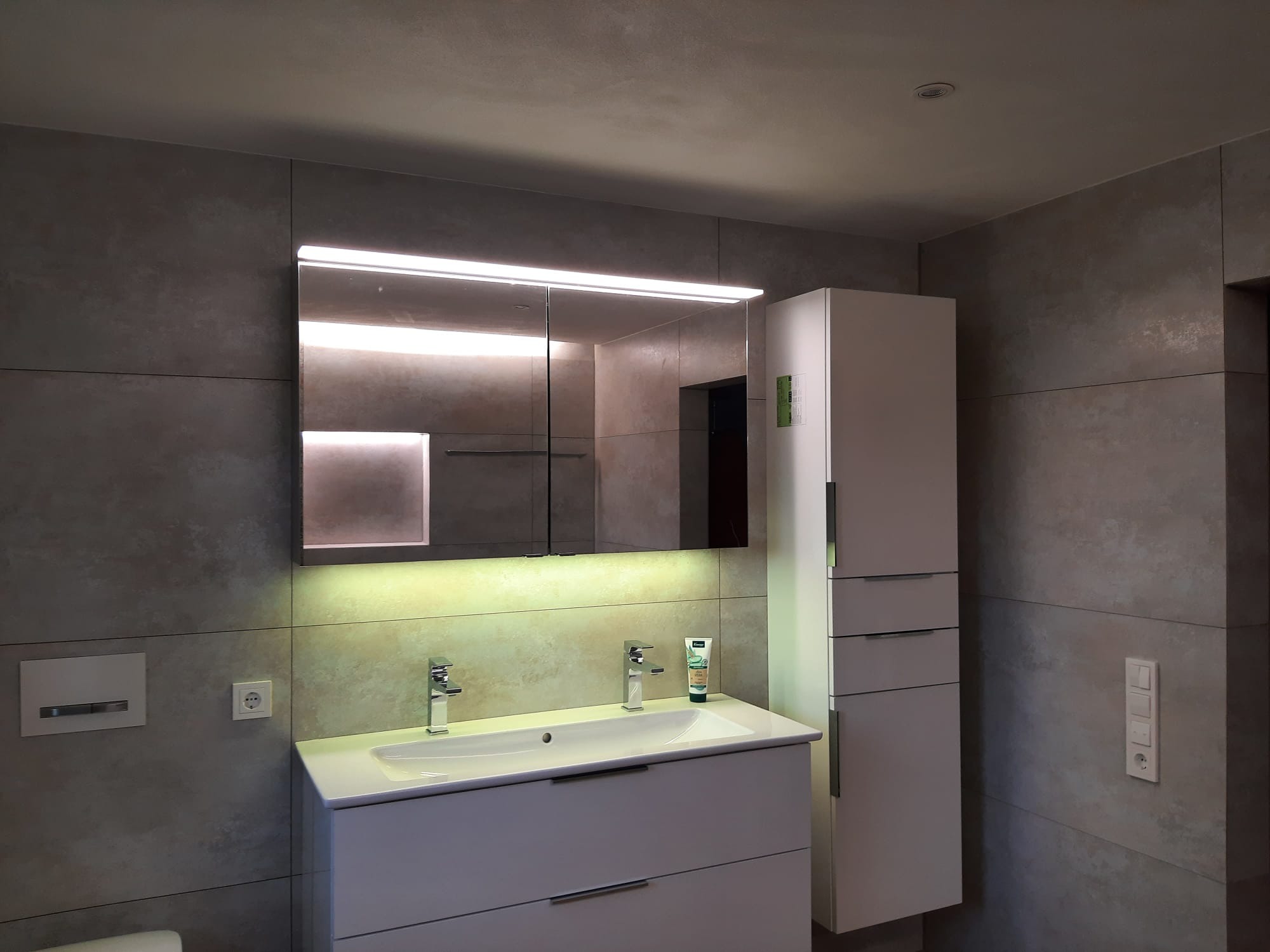 Nowoczesna łazienka z szarymi płytkami, białą szafką pod umywalką i lustrem z oświetleniem LED. Minimalistyczny design z akcentami chromu i subtelnym oświetleniem.