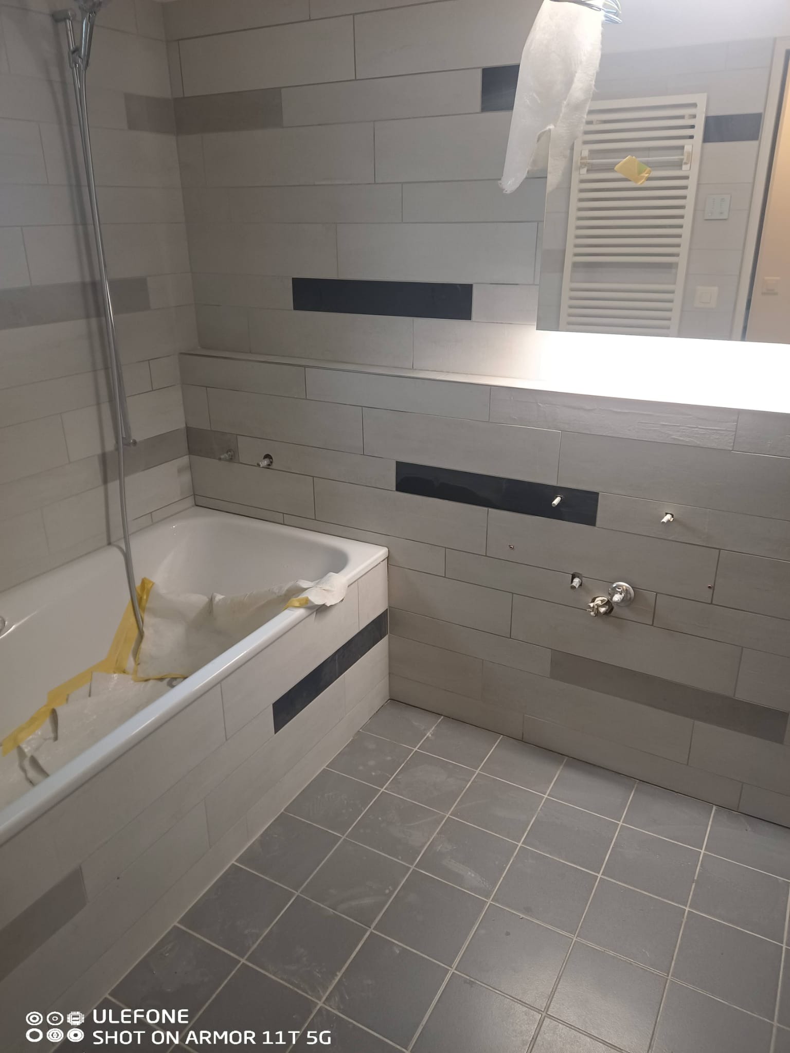 Wykończona łazienka z wanną, szarą glazurą w poziome pasy z czarnymi akcentami, szarą podłogą i lustrem z oświetleniem w tle. Widoczne elementy instalacji.