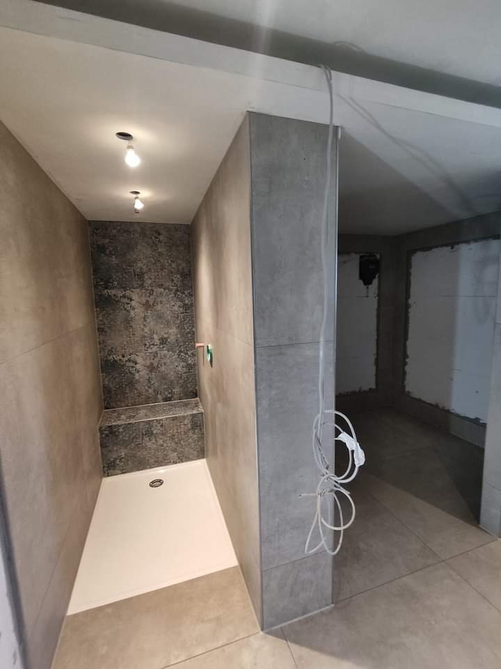 Nowoczesna łazienka w szarościach z kabiną typu walk-in i siedziskiem, oświetlona punktowo. Widoczne niedokończone prace w sąsiednim pomieszczeniu.