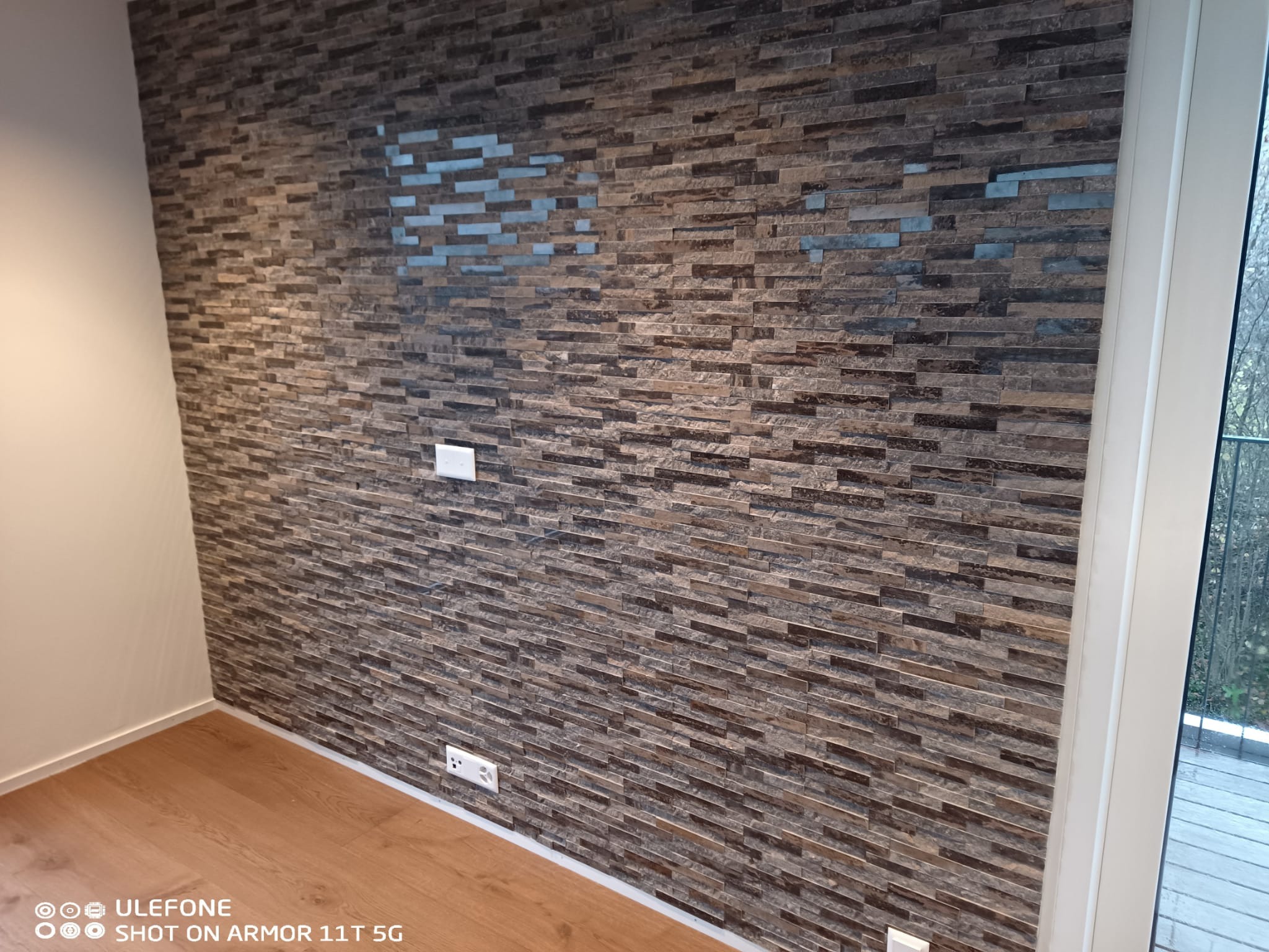 Ściana wykończona dekoracyjną, brązowo-szarą okładziną imitującą kamień, z widocznymi gniazdkami elektrycznymi i fragmentem podłogi z paneli.
