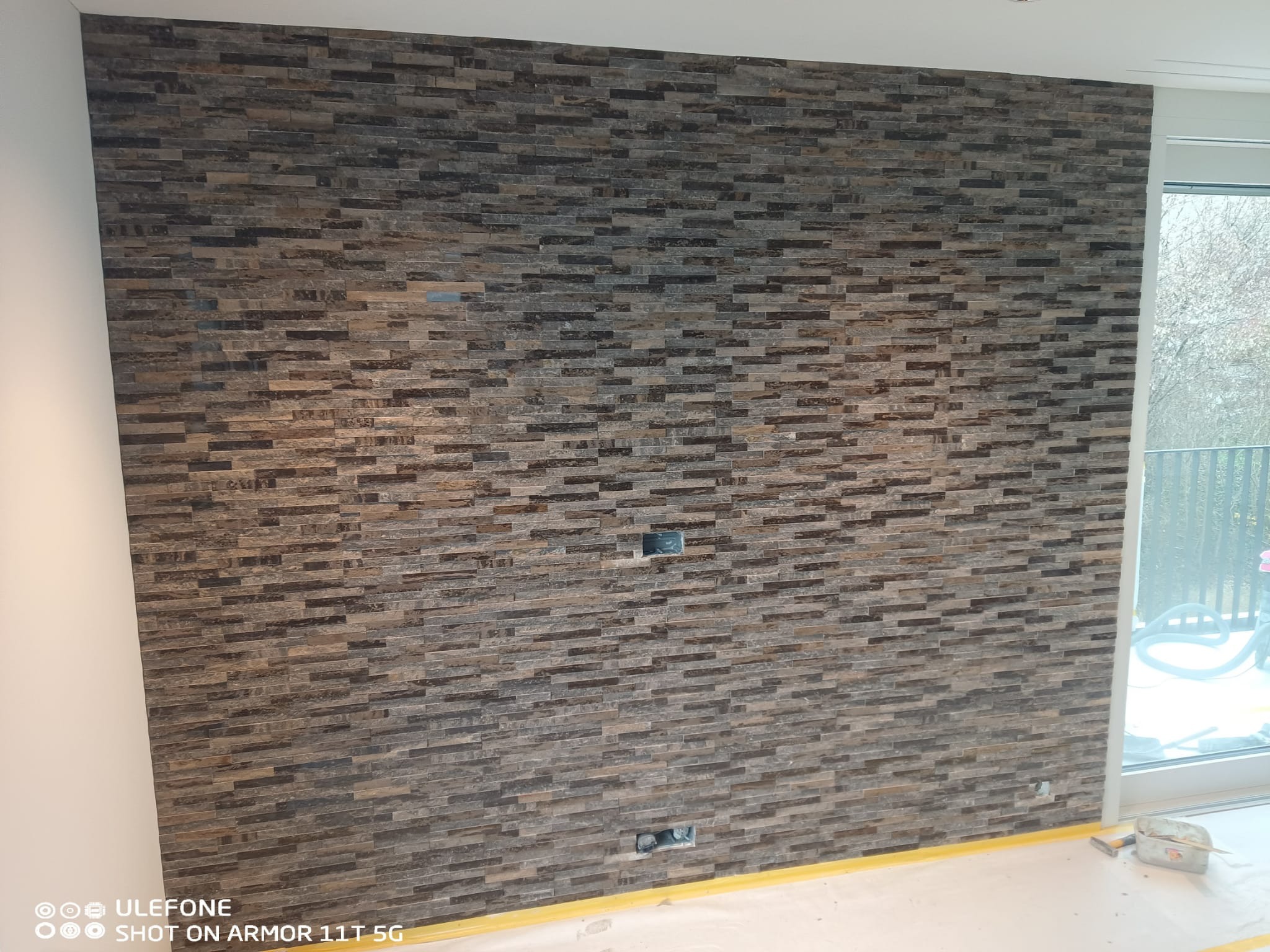 Ściana wykończona dekoracyjną, brązowo-szarą okładziną z małych, prostokątnych płytek, z widocznymi gniazdkami elektrycznymi i fragmentem okna.