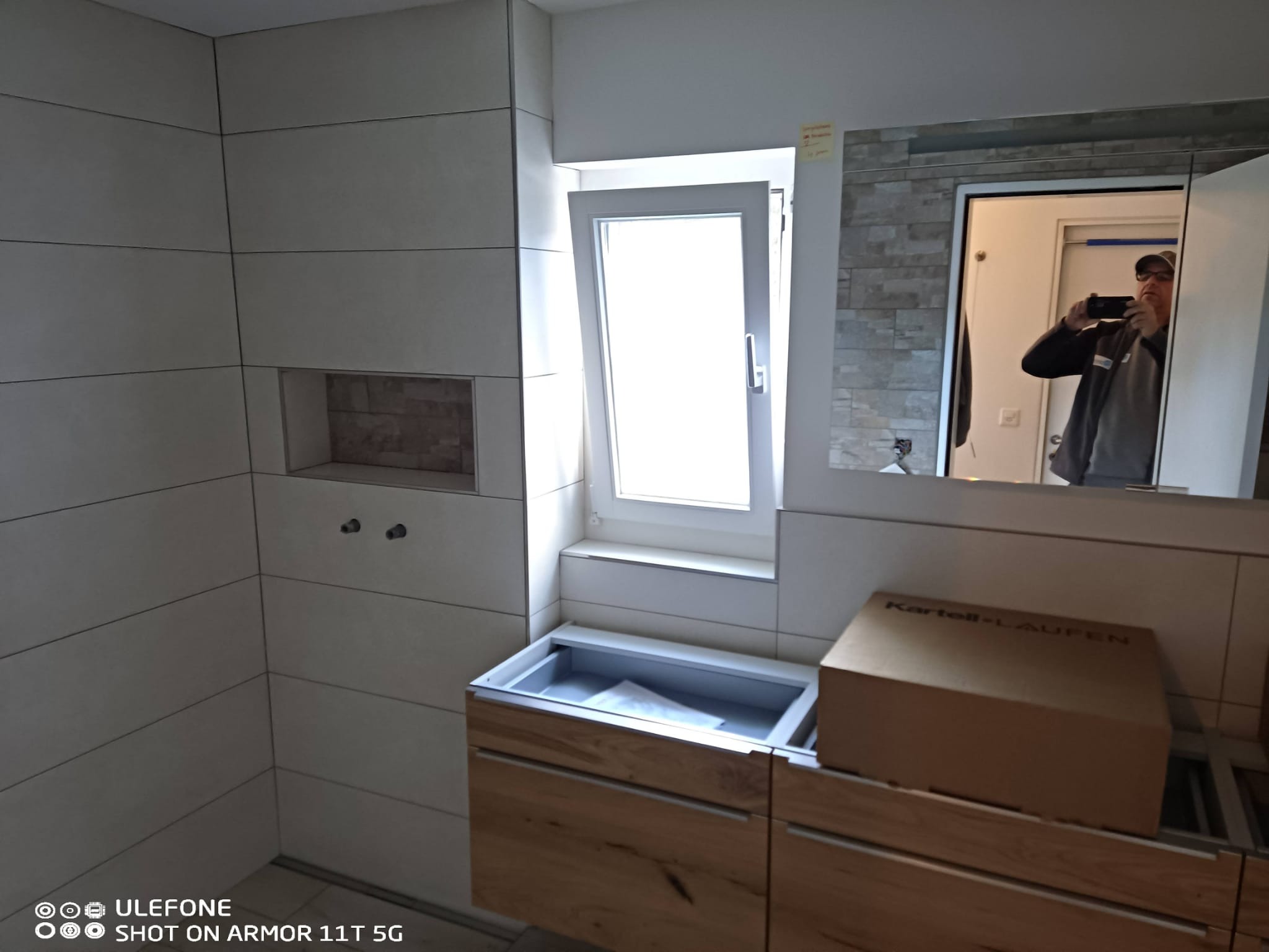 Wykończona łazienka z beżowymi płytkami, drewnianą szafką pod umywalkę i oknem. W lustrze odbija się osoba robiąca zdjęcie. Widoczny karton z napisem Kartell-Laufen.