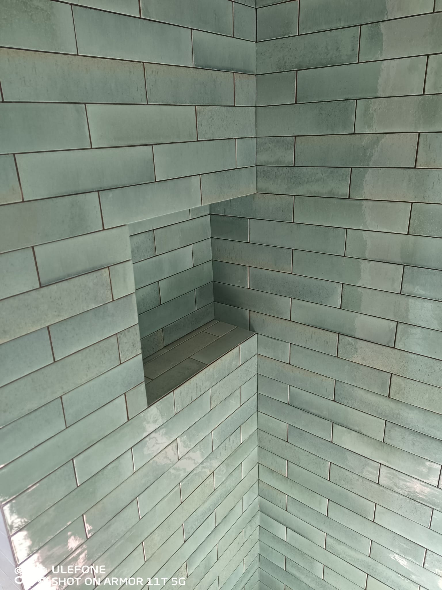 Narożnik wykończony podłużnymi, turkusowymi płytkami z wnęką. Estetyczne połączenie płytek, widoczne fugi. Wykończenie w łazience.