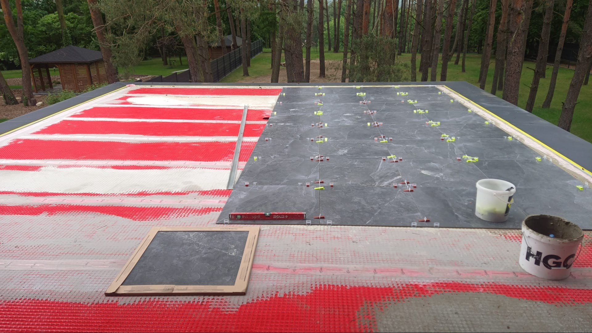 Układanie ciemnych płytek na balkonie, widoczna hydroizolacja w czerwono-białe pasy, poziomica i wiaderka z zaprawą. Prace w toku, tło: drzewa.