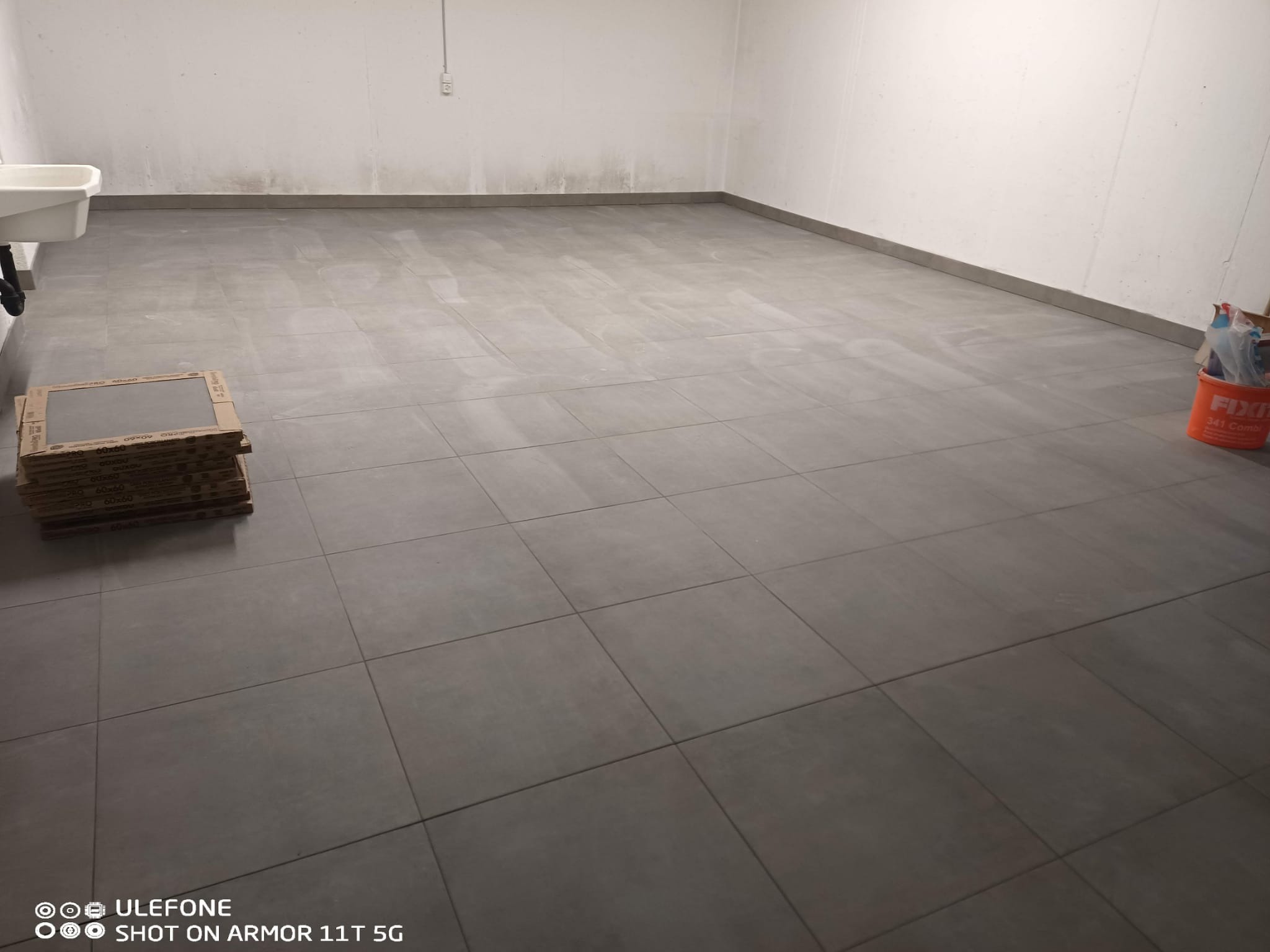 Ułożona szara terakota podłogowa w surowym pomieszczeniu. Na podłodze stos płytek i wiadro z klejem. Widoczny fragment umywalki.