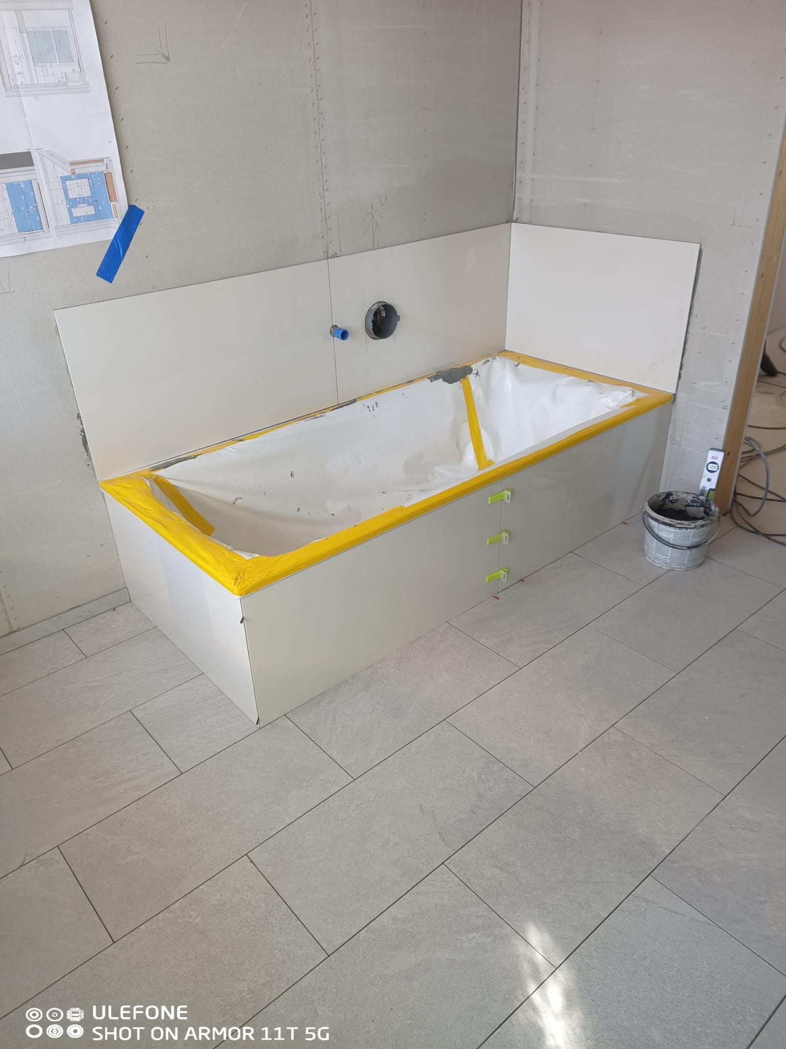 Wanna w obudowie z płyt, zabezpieczona żółtą taśmą, z zielonymi krzyżykami dystansowymi. Na podłodze szare płytki. Na ścianie widoczny projekt łazienki.