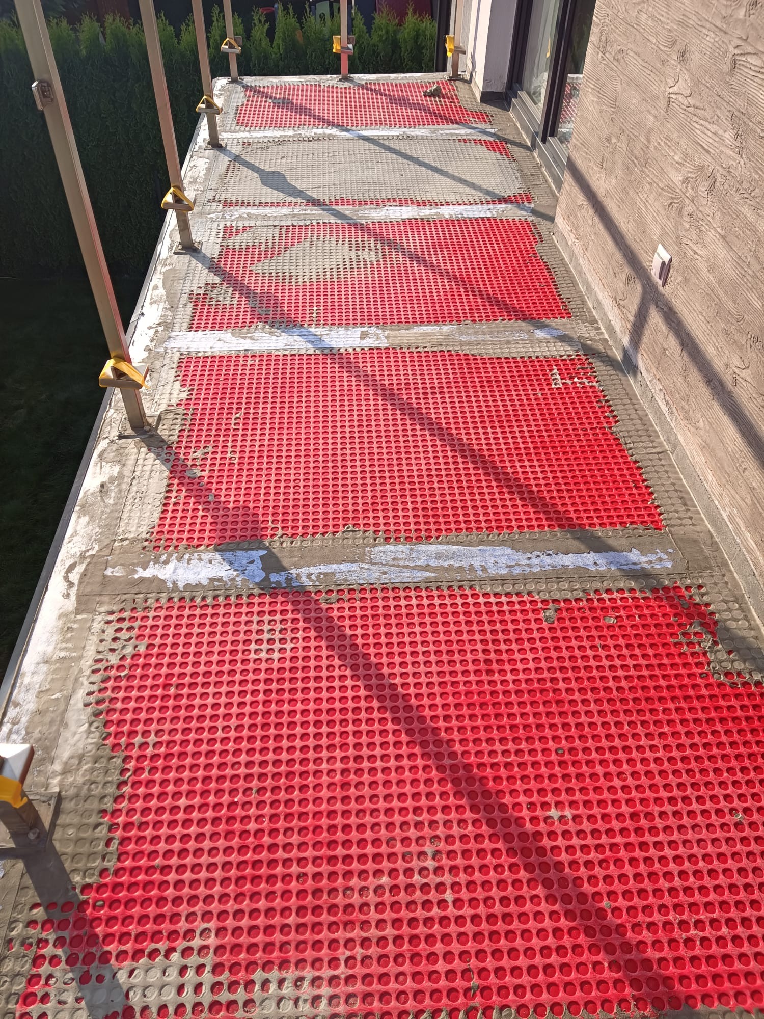 Balkon w trakcie prac: czerwona mata drenażowa, częściowo zalana zaprawą, przygotowana pod wylewkę. Widoczne rusztowanie i elewacja budynku.