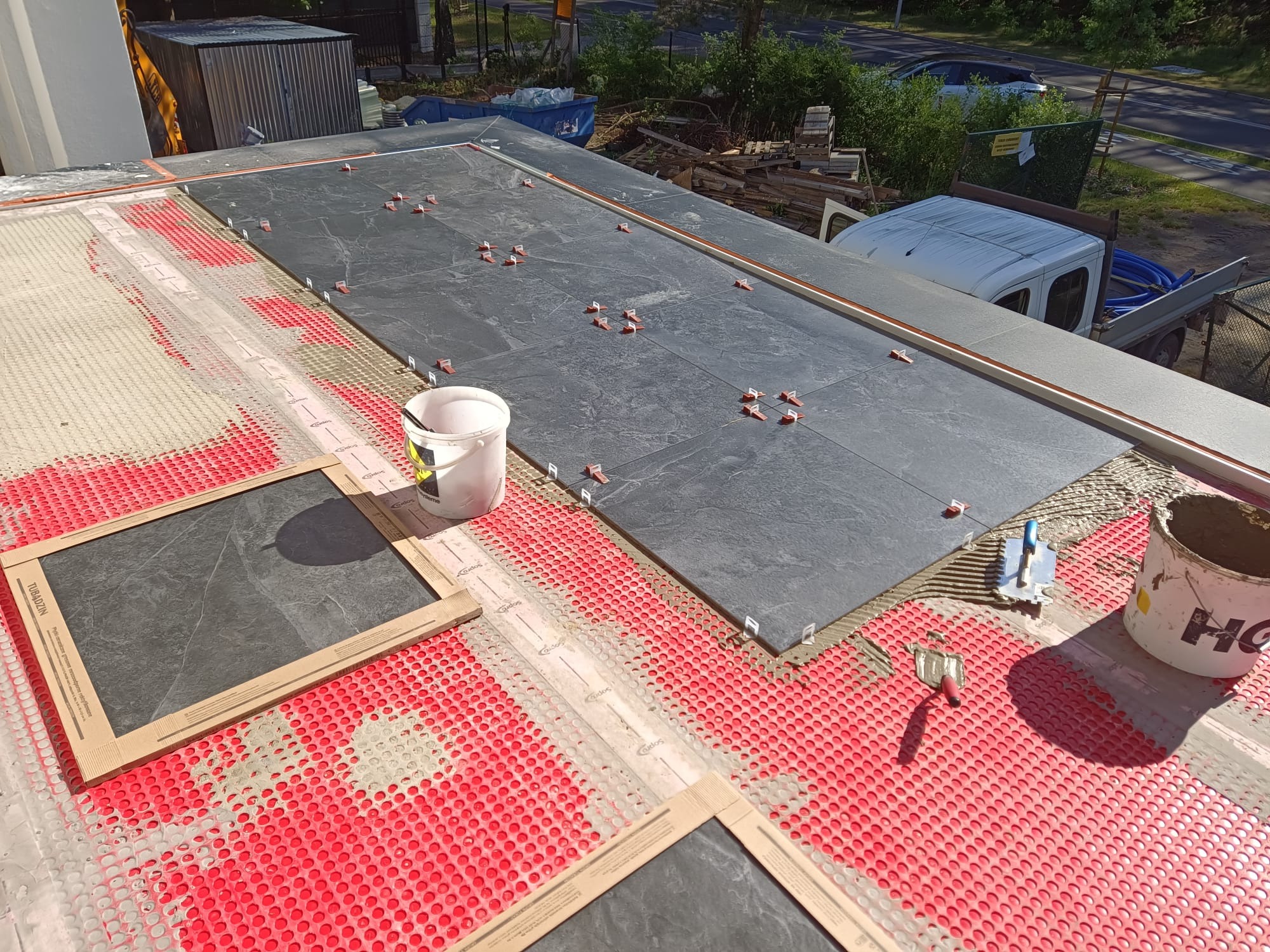 Układanie ciemnych płytek na czerwonej macie drenażowej, widoczne narzędzia glazurnicze i wiaderka z zaprawą. Prace na balkonie lub tarasie w słoneczny dzień.