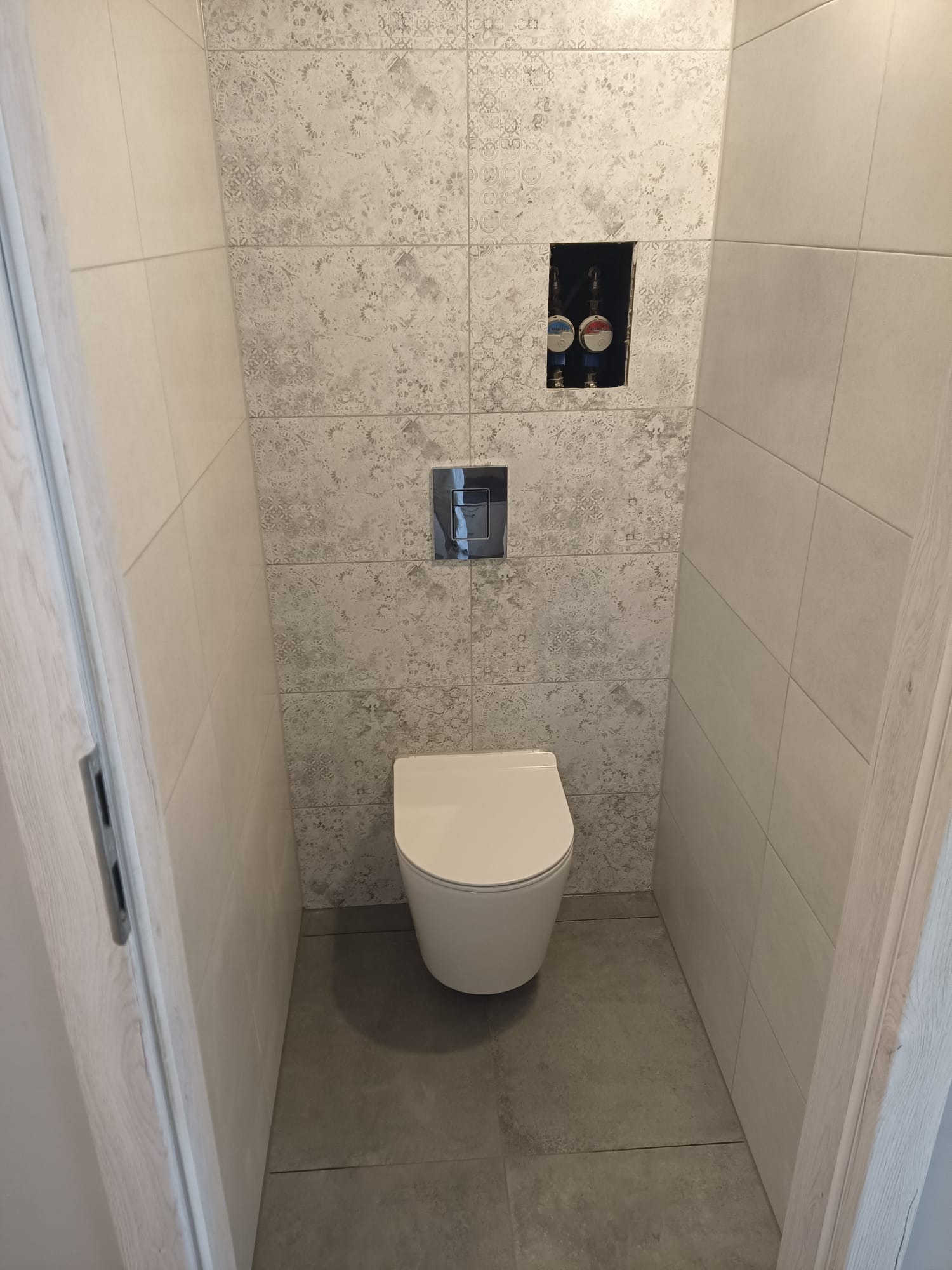 Kompakt WC z podtynkową spłuczką na tle ściany z szarych płytek z delikatnym wzorem. Widoczne zawory i chromowany przycisk spłuczki. Minimalistyczny design łazienki.