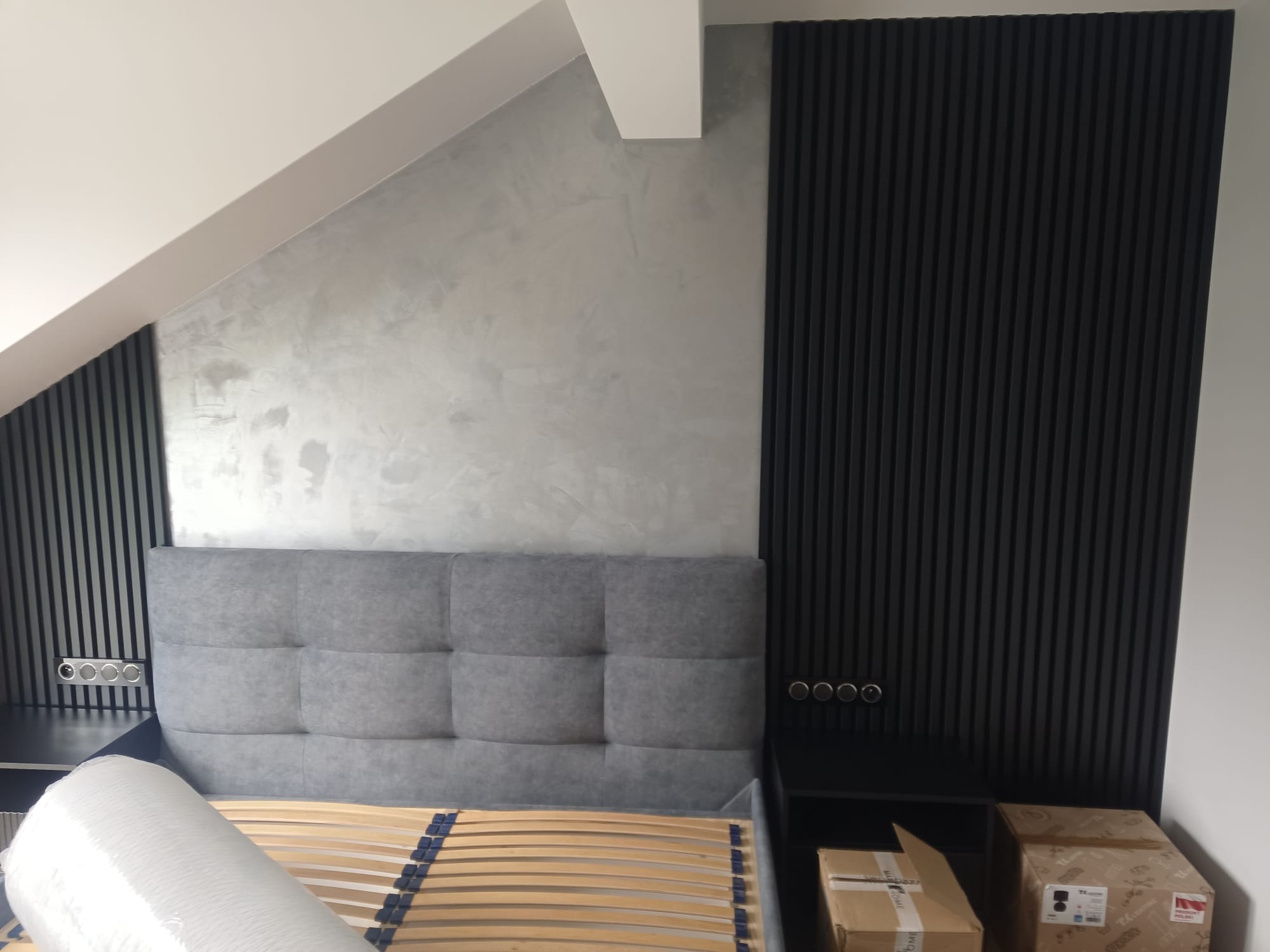 Szara sypialnia na poddaszu: tapeta strukturalna, czarne lamele na ścianie za wezgłowiem łóżka. Na szafce nocnej kartony, widoczny stelaż łóżka.