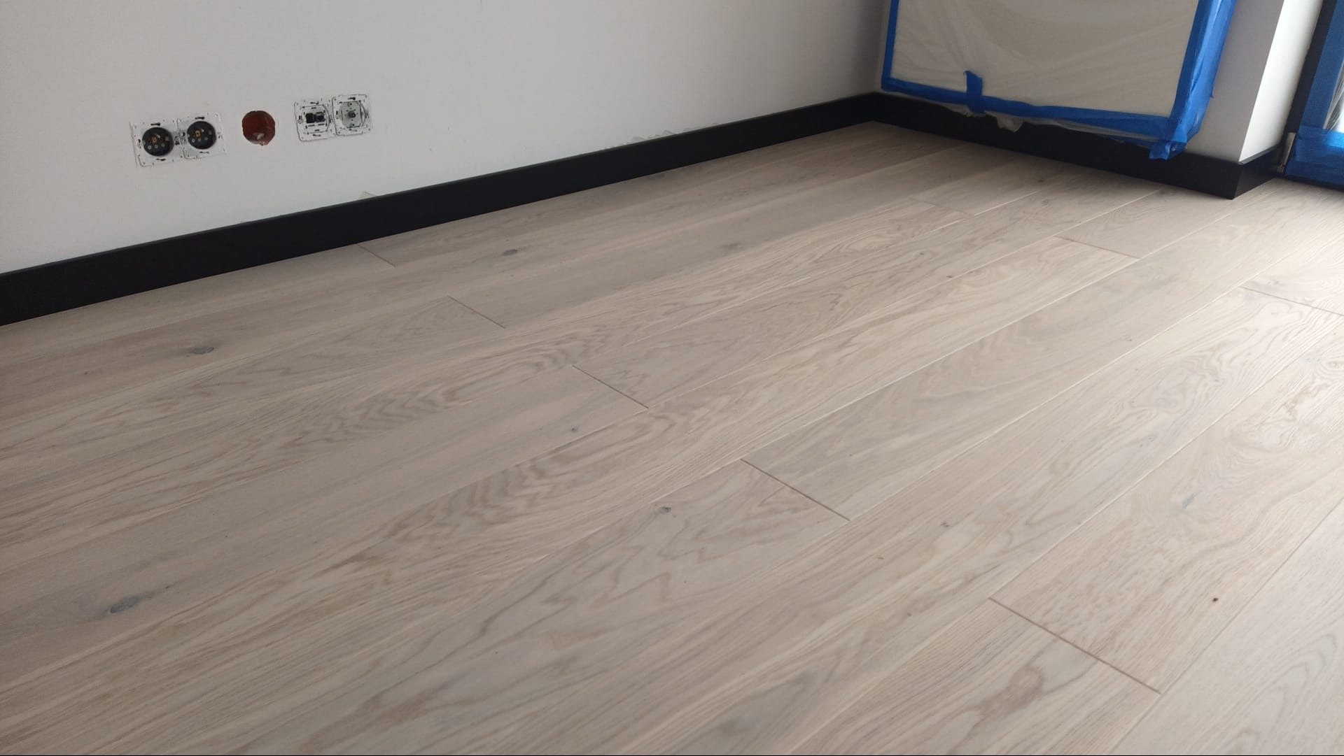 Jasne panele podłogowe ułożone w pokoju z czarnymi listwami przypodłogowymi i gniazdkami elektrycznymi w ścianie. Fragment zabezpieczonego okna.