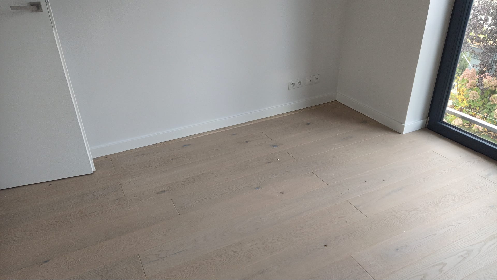 Jasne panele podłogowe w pokoju z białymi ścianami, listwami przypodłogowymi i oknem balkonowym. Widoczne gniazdka elektryczne.