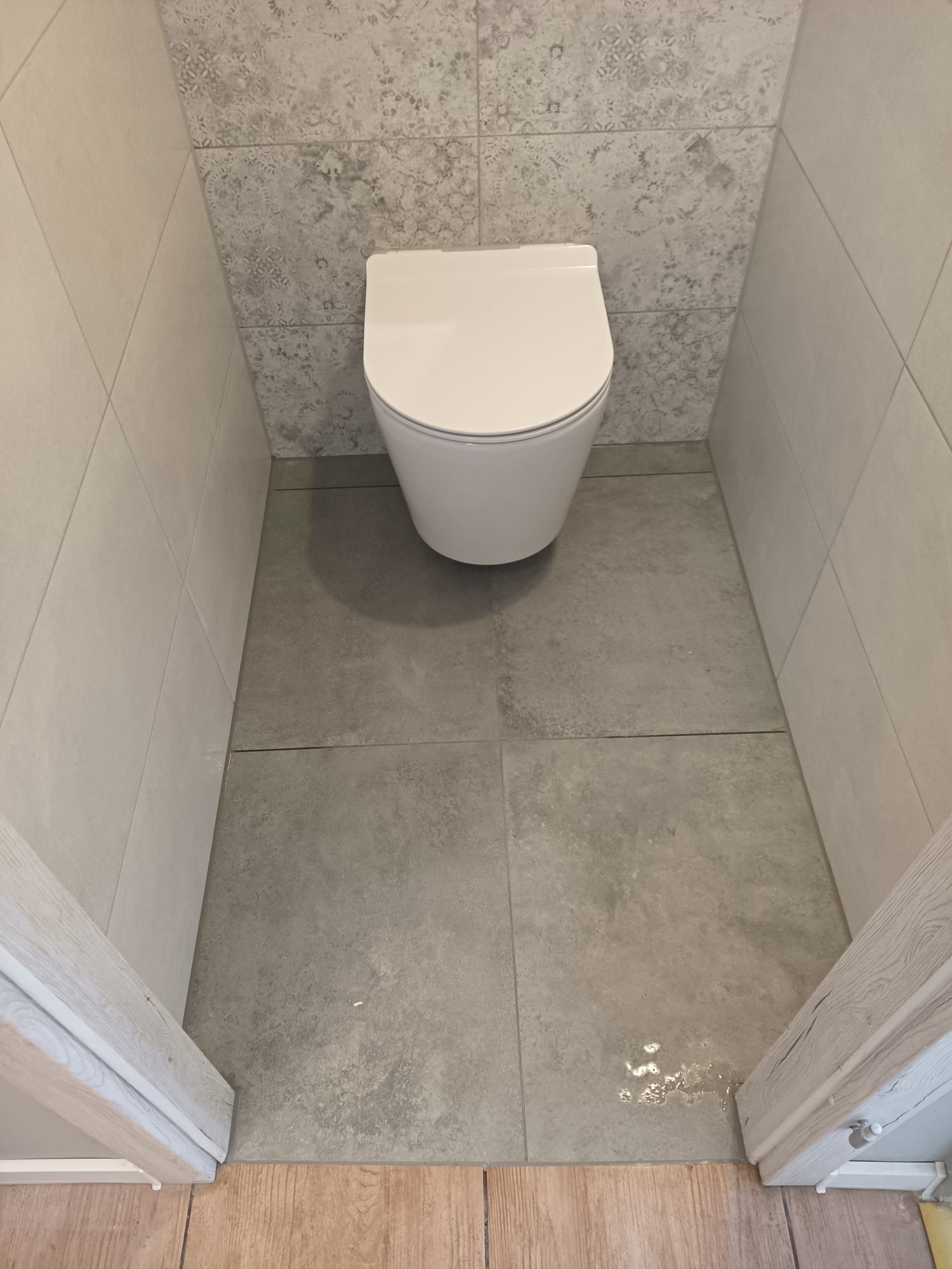 Biała, podwieszana toaleta w wykończonej łazience. Szare płytki podłogowe i dekoracyjne płytki ścienne. Fragment drewnianej ramy drzwi w dolnej części kadru.