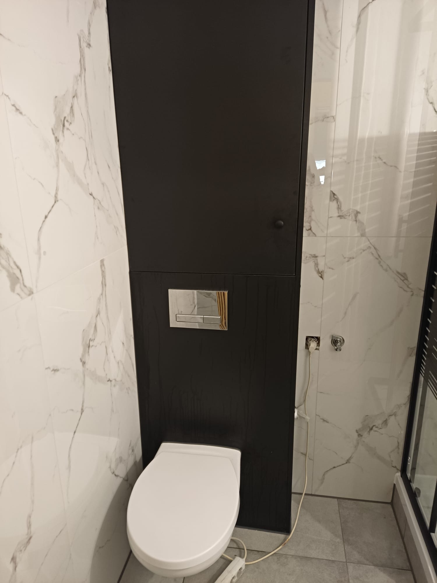 Nowoczesna toaleta podwieszana z czarną zabudową i marmurowymi płytkami. Widoczny fragment kabiny prysznicowej i gniazdko elektryczne.