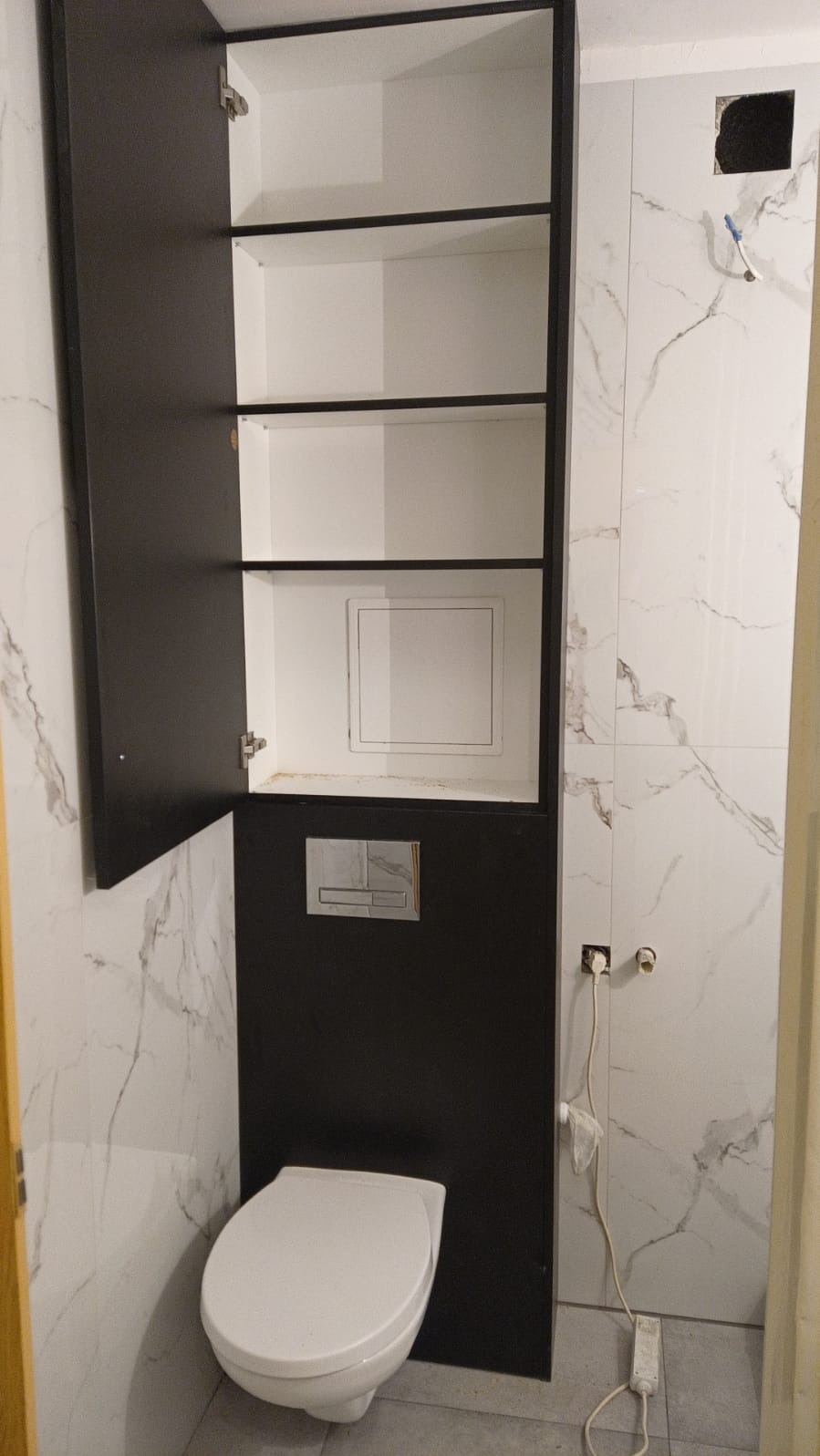Wykończona łazienka z wiszącą toaletą, zabudową szafkową w czerni i bieli oraz marmurowymi płytkami. Widoczne gniazdka elektryczne i otwarta szafka.
