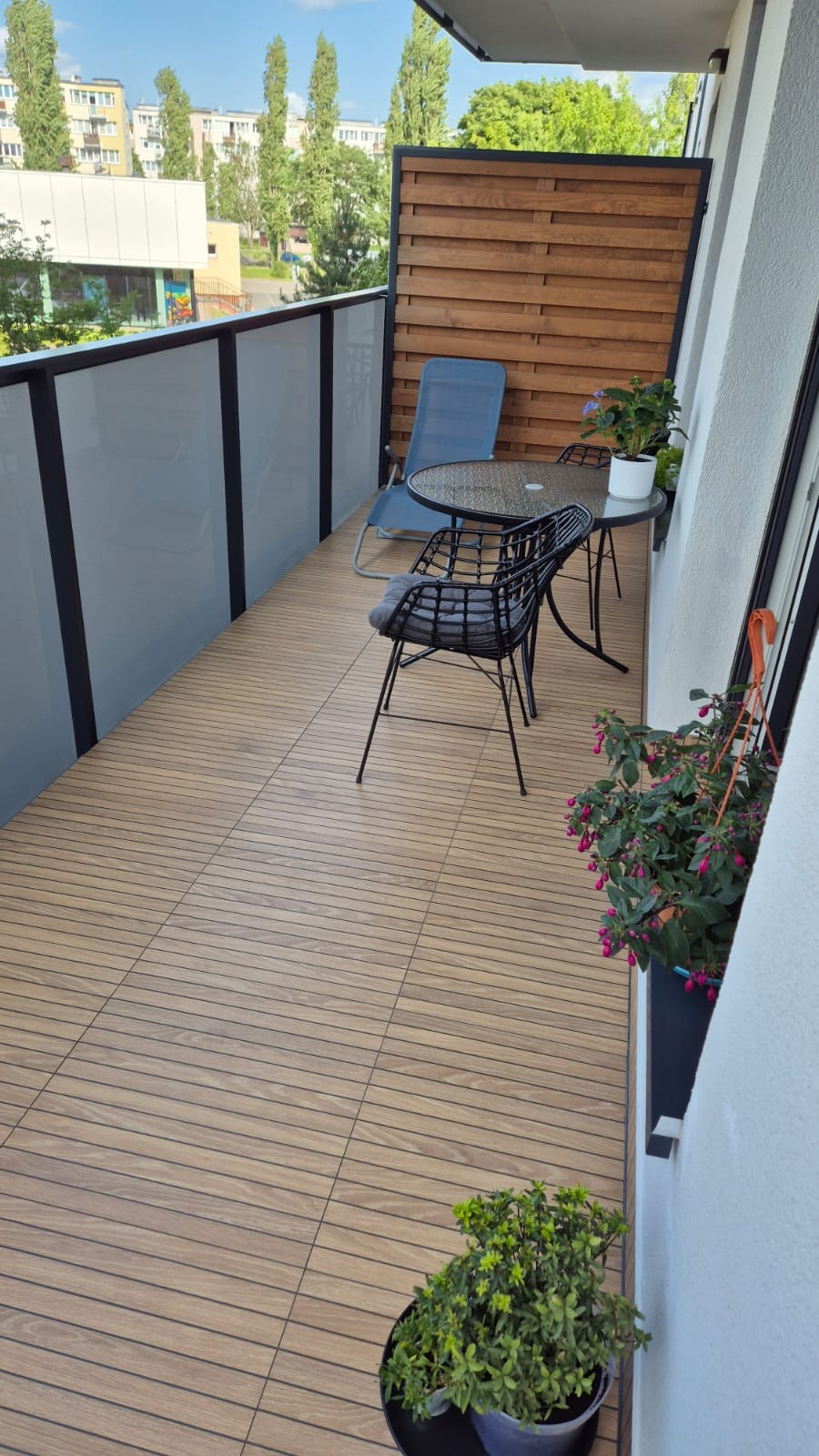 Balkon z drewnopodobną podłogą z płytek, stolikiem i krzesłami, ozdobiony zielenią w doniczkach. Widok na zewnątrz przez przeszkloną balustradę.