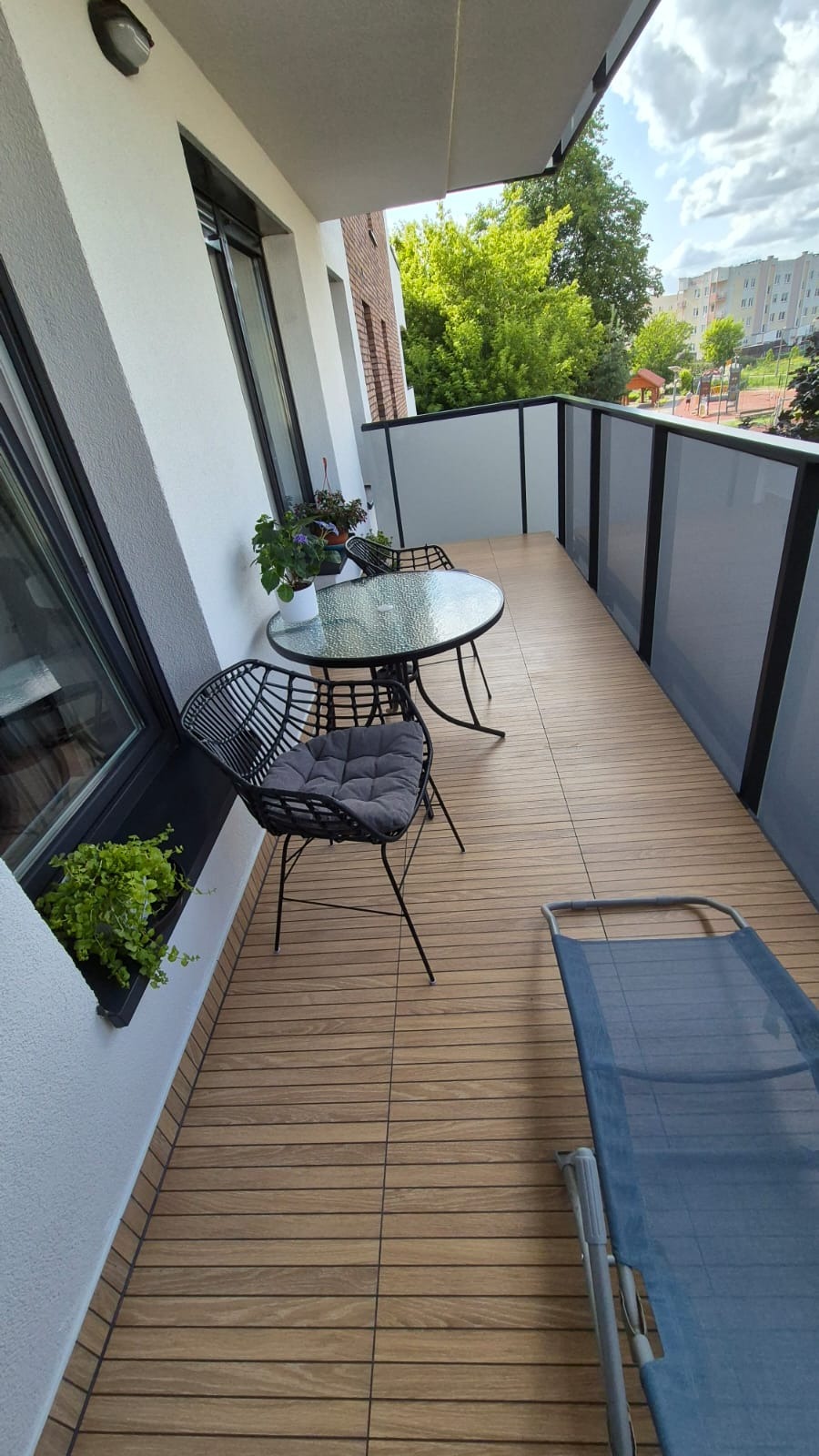 Balkon z drewnopodobną terakotą, stolikiem kawowym i krzesłami. Widok na zieleń i plac zabaw w tle. Nowoczesne balustrady z szarego szkła.