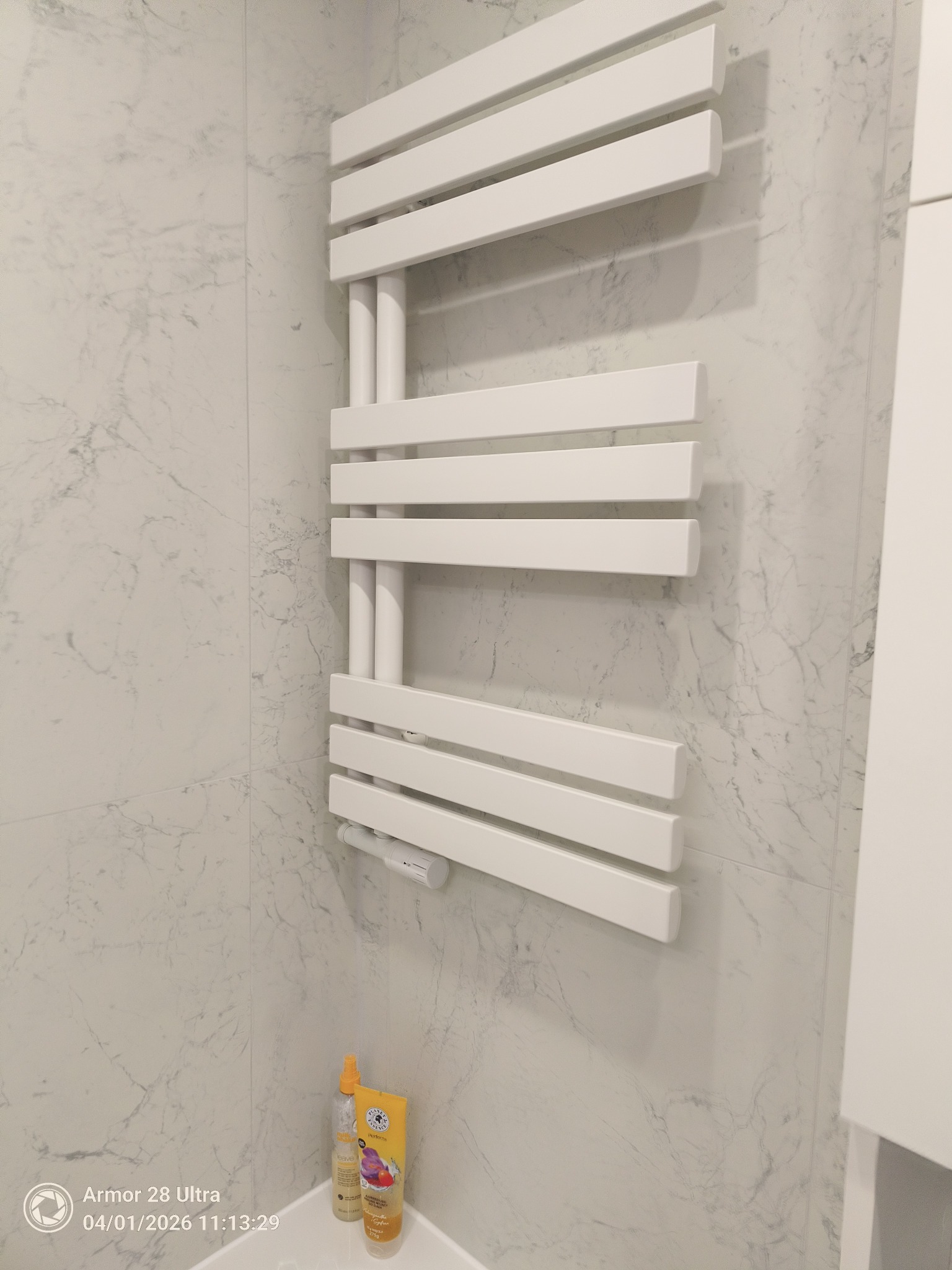 Biały, drabinkowy grzejnik łazienkowy na ścianie z płytek imitujących marmur. Widoczne kosmetyki na umywalce. Minimalistyczny design, nowoczesne wykończenie łazienki.