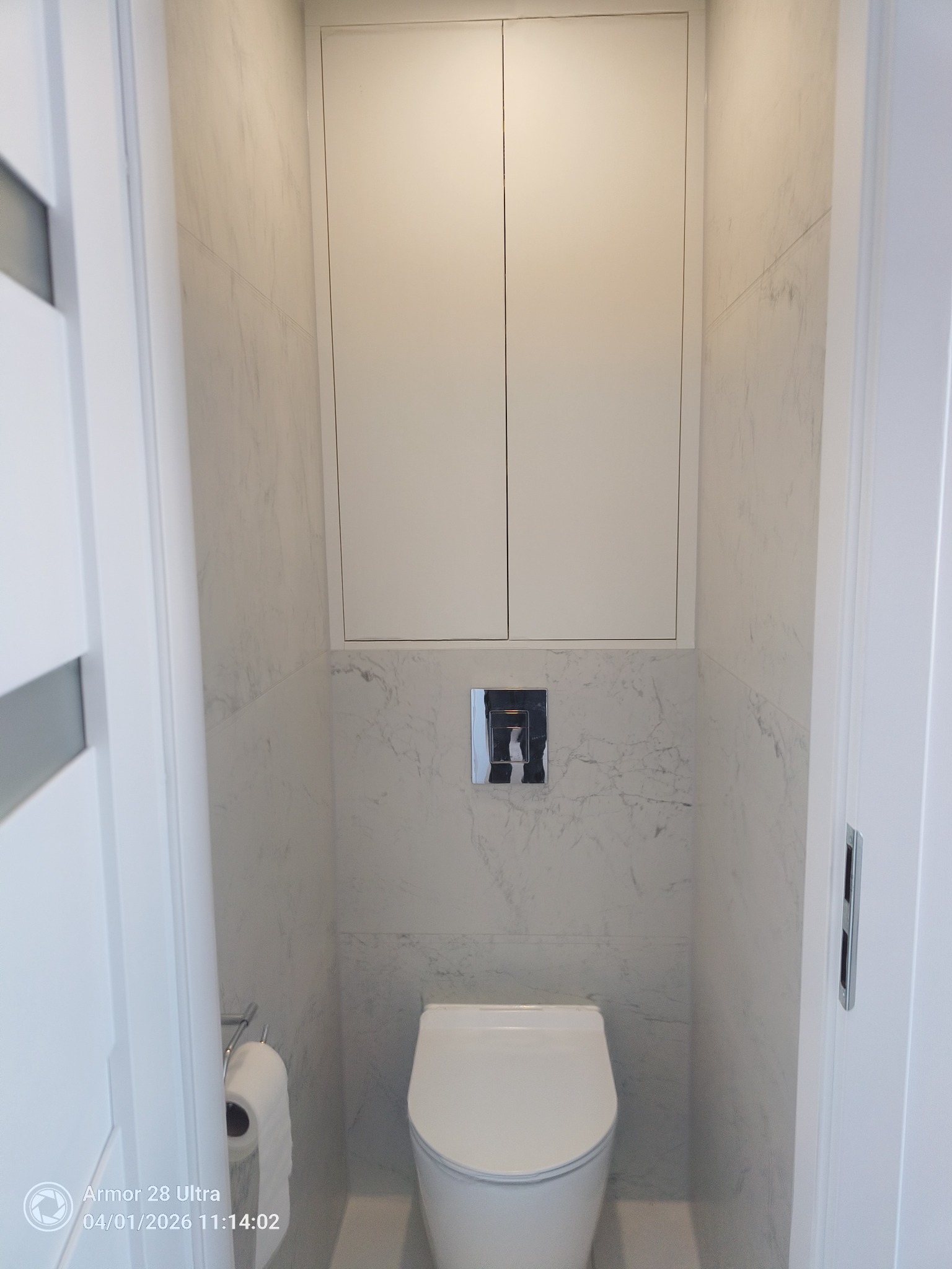 Nowoczesna toaleta w małej łazience wykończonej białymi płytkami imitującymi marmur, z zabudową nad stelażem i minimalistycznym przyciskiem spłukującym.