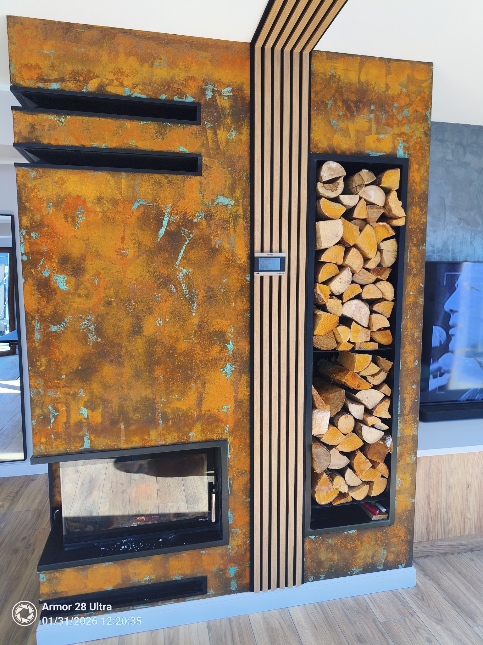 Rustykalny kominek z wnęką na drewno, wykończony metaliczną farbą z efektem rdzy i drewnianymi listwami. Nowoczesny design z elementami industrialnymi.