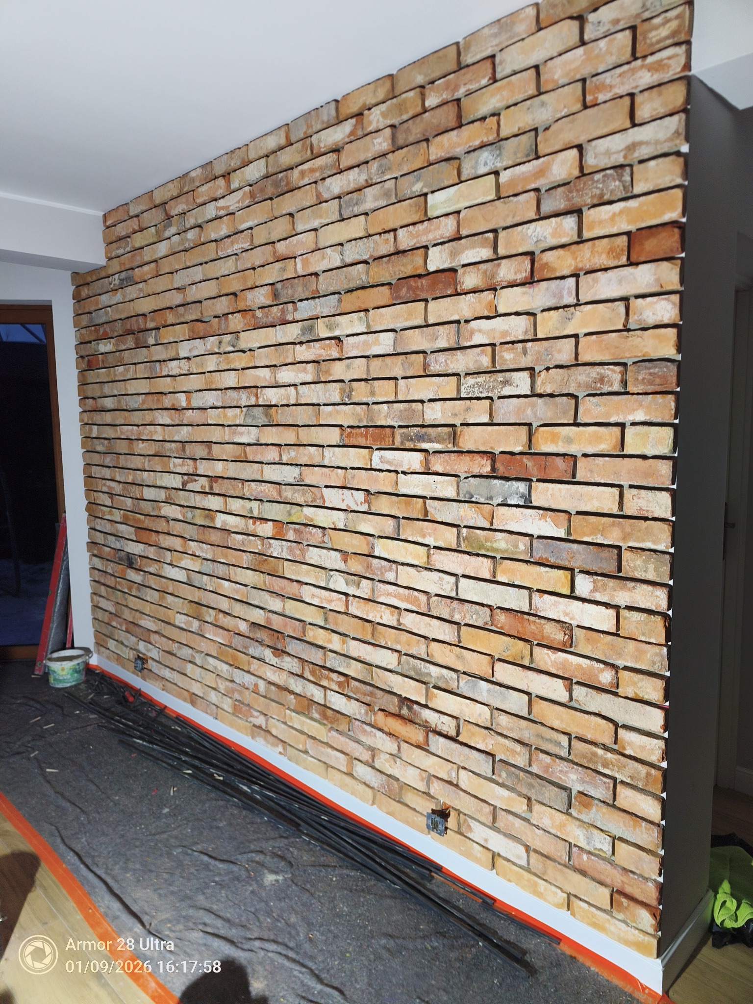 Ściana wyłożona dekoracyjną cegłą w odcieniach beżu i brązu, widoczna z bliska, z elementami przygotowania do dalszych prac wykończeniowych.