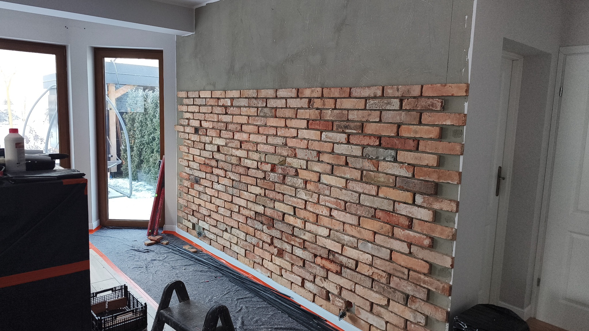 Ceglana ściana dekoracyjna w trakcie montażu, widoczne narzędzia i przygotowane podłoże. Częściowo wykończona ściana z cegły w pomieszczeniu.