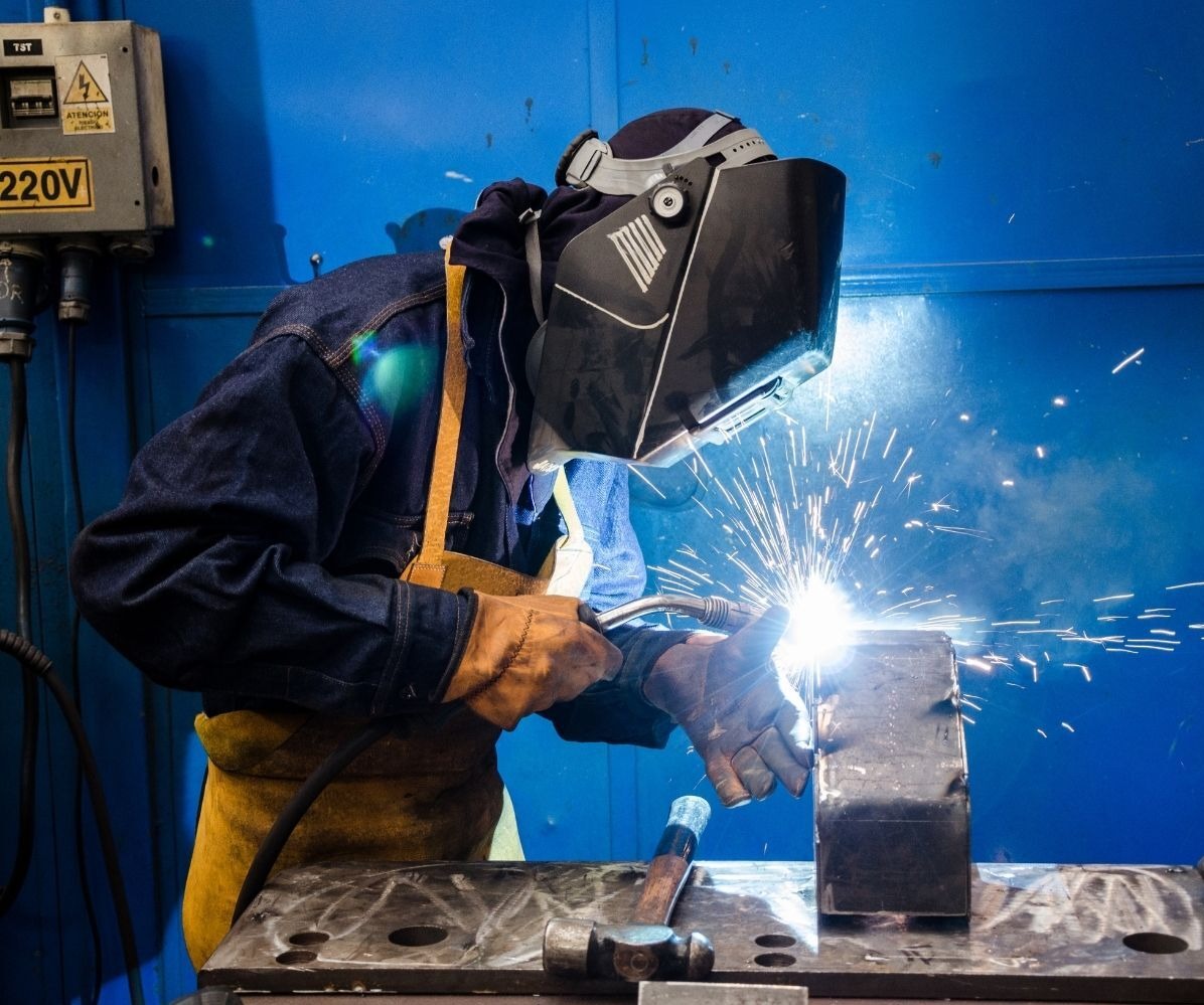 Spawanie metalu w Bytowie: spawacz w masce, iskry i dym podczas pracy nad konstrukcją. Widoczny młotek i stanowisko robocze na tle niebieskiej ściany.