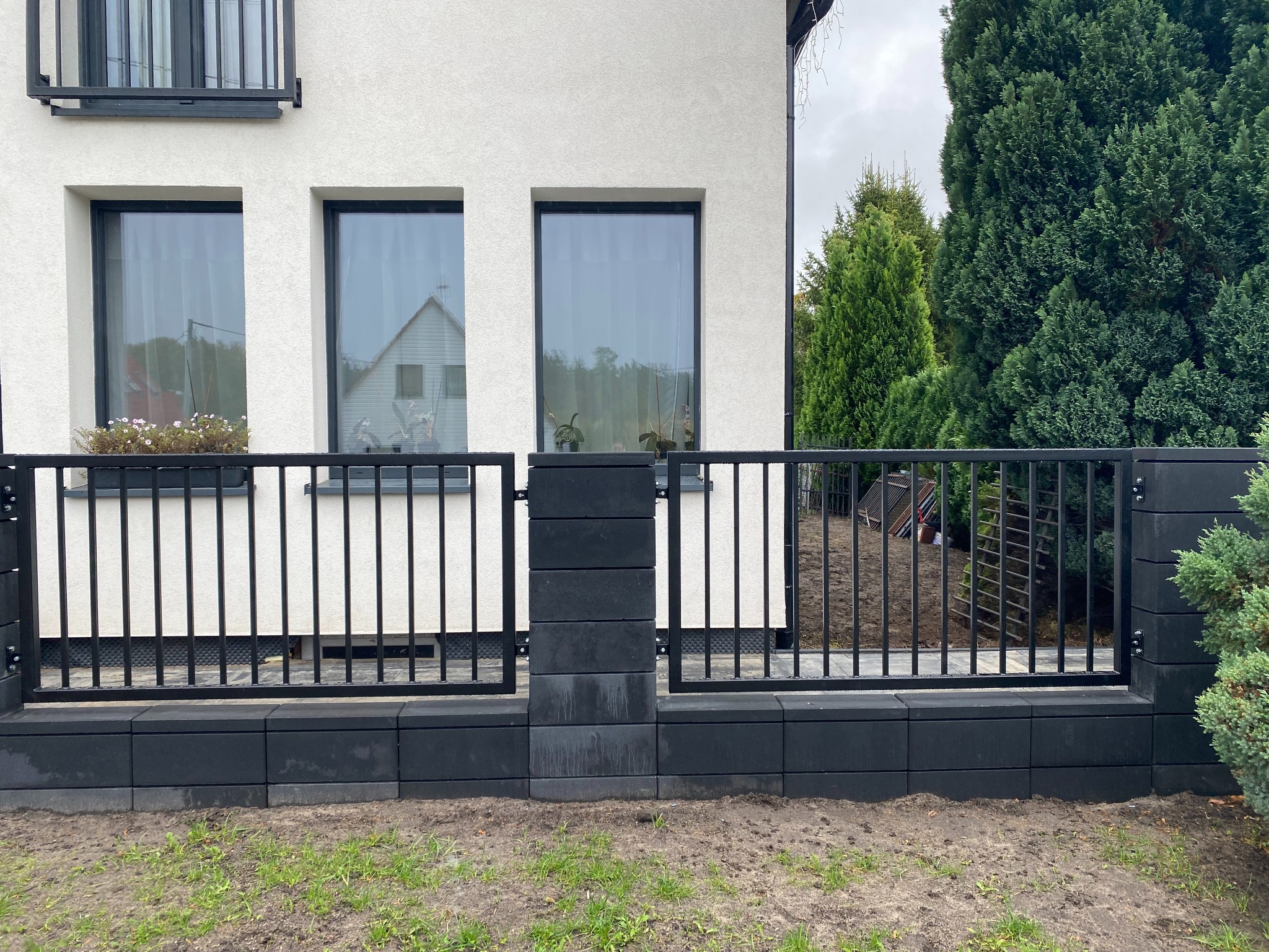 Nowoczesne ogrodzenie z czarnych, prostych elementów, na tle białego domu z dużymi oknami. Minimalistyczny design i staranne wykonanie.